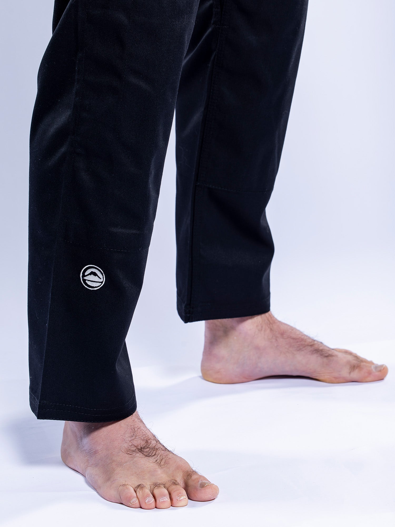 A person stands barefoot on a white surface, wearing the Baseline 2.0 BJJ Gi Black Grey pants featuring a small circular logo near the left ankle.