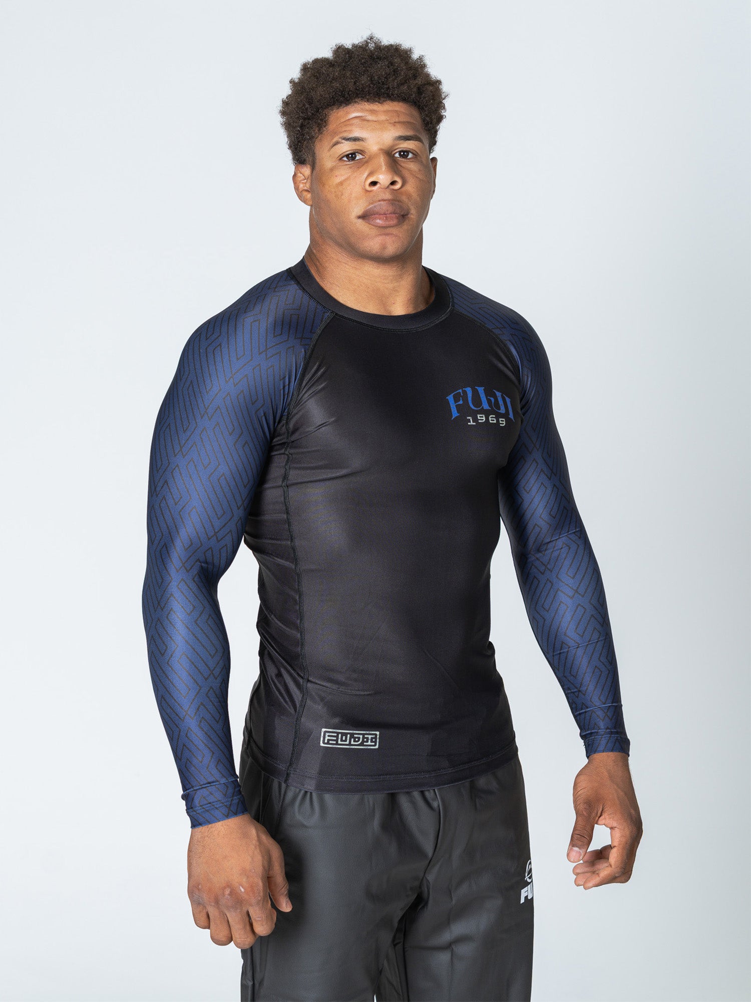 A muscular man wears the Oni Samurai Flex Lite Rashguard Blue with black shorts, standing against a plain light background and looking directly at the camera with a neutral expression.