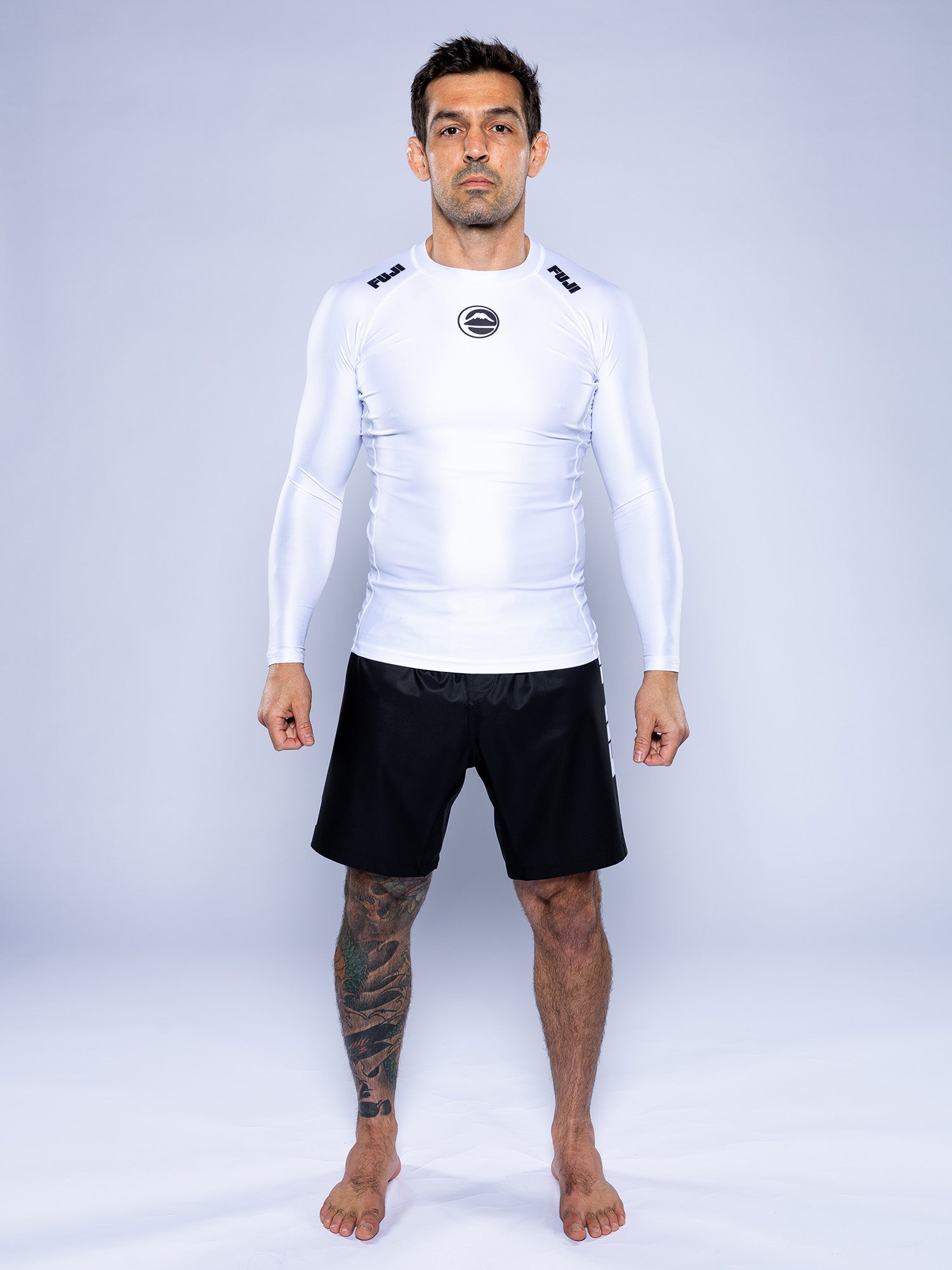 A man with short dark hair and a left leg tattoo stands barefoot on a white background, wearing the Baseline 2.0 Long Sleeve Rashguard in white and black shorts.