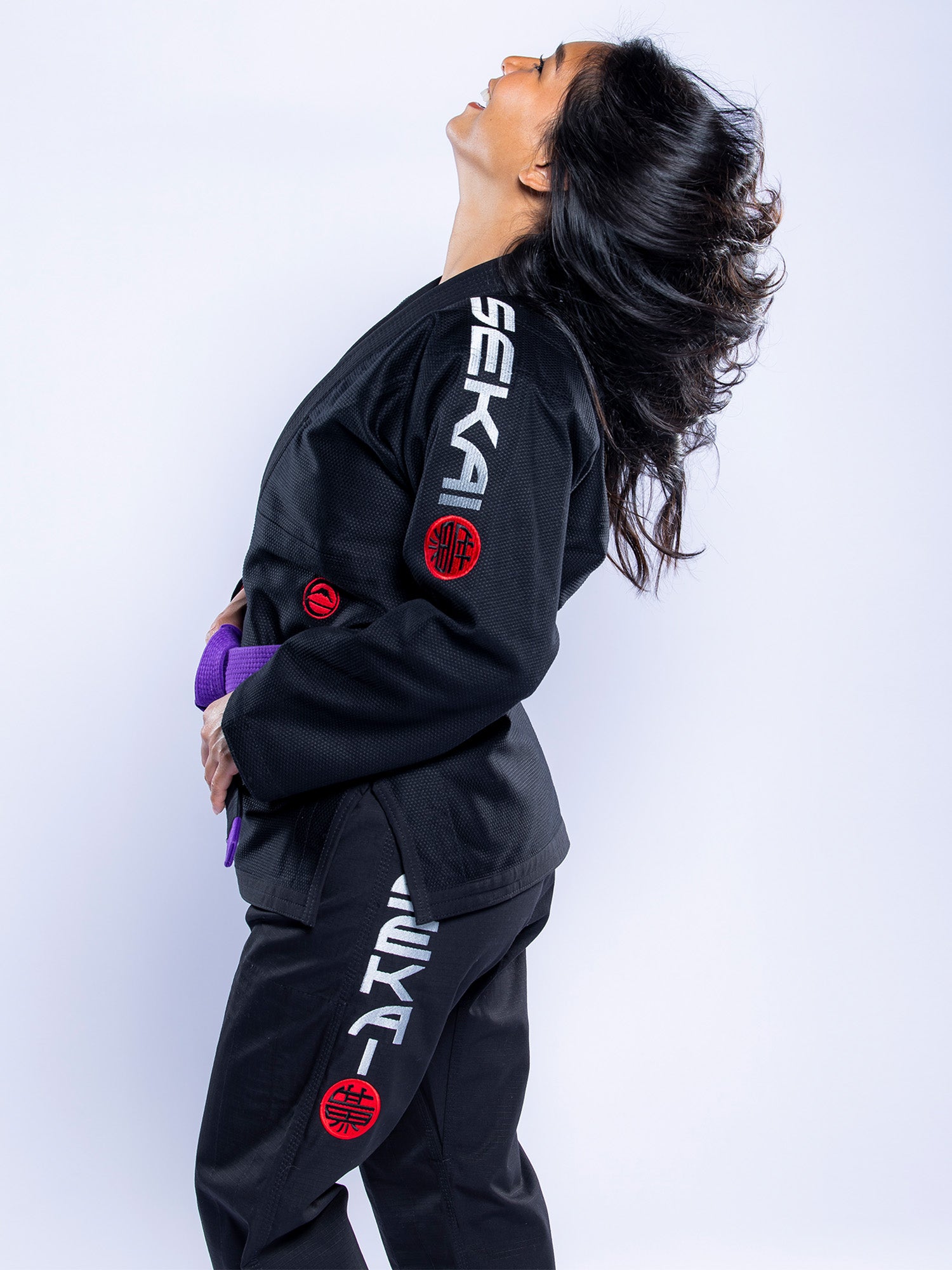 A woman in the Womens SEKAI 3.0 BJJ Gi Black Grey stands in profile, holding a purple belt and tossing her hair back, with "SEKAI" text and red patches visible on the gi.