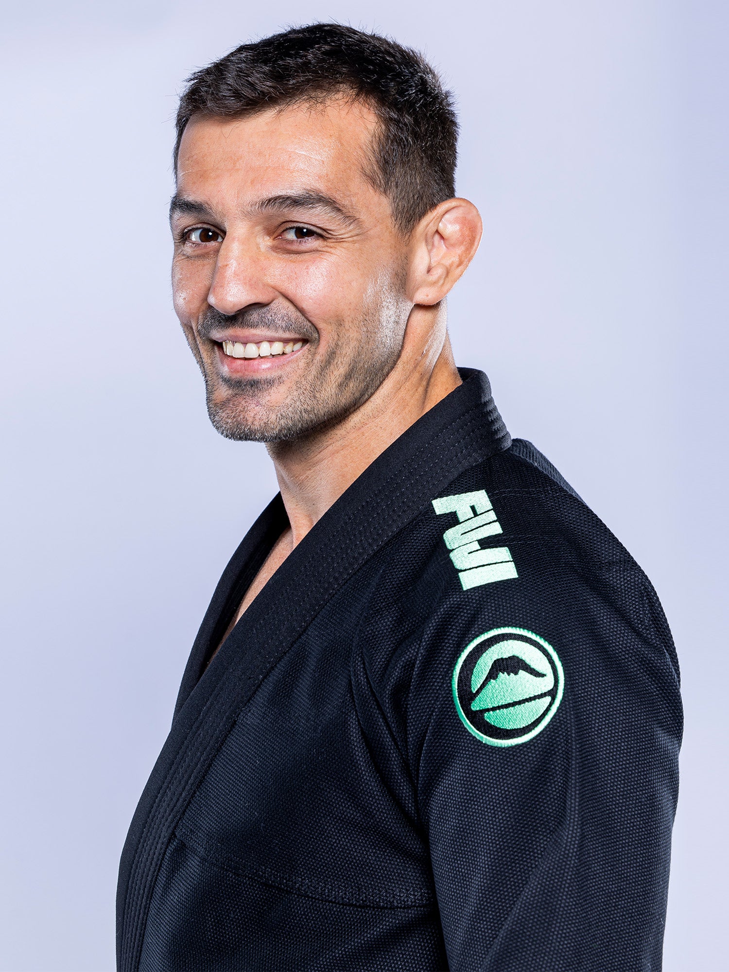 A man with short dark hair and a trimmed beard smiles, wearing the Baseline 2.0 BJJ Gi Black Mint, featuring FUJI branding and a green mountain logo on the sleeve, standing against a light background.