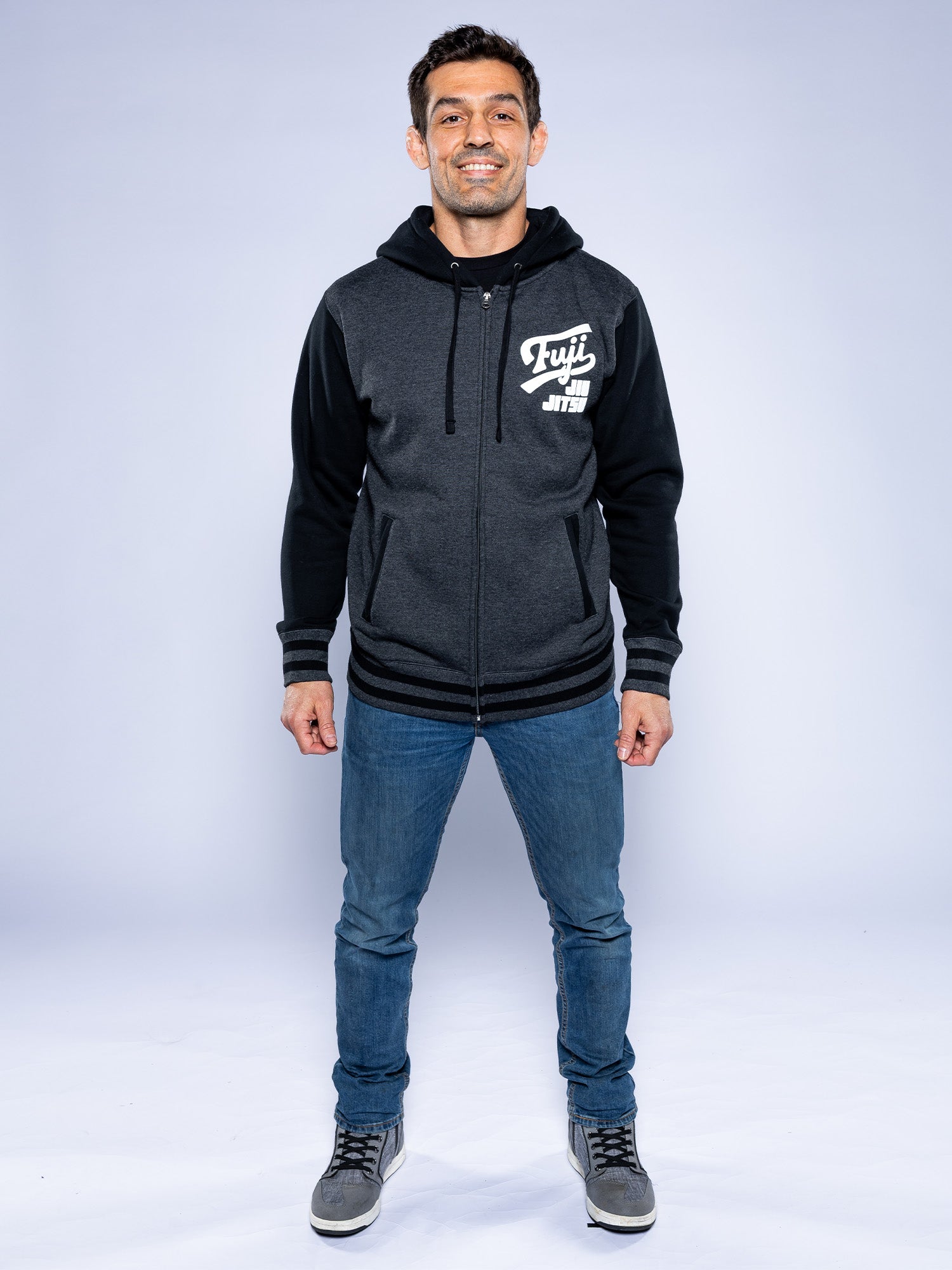 A man smiles, wearing the Jiu Jitsu Varsity Zip Hoodie in charcoal with “Fuji Jiu Jitsu” on the chest, blue jeans, and gray sneakers, standing against a plain light background.