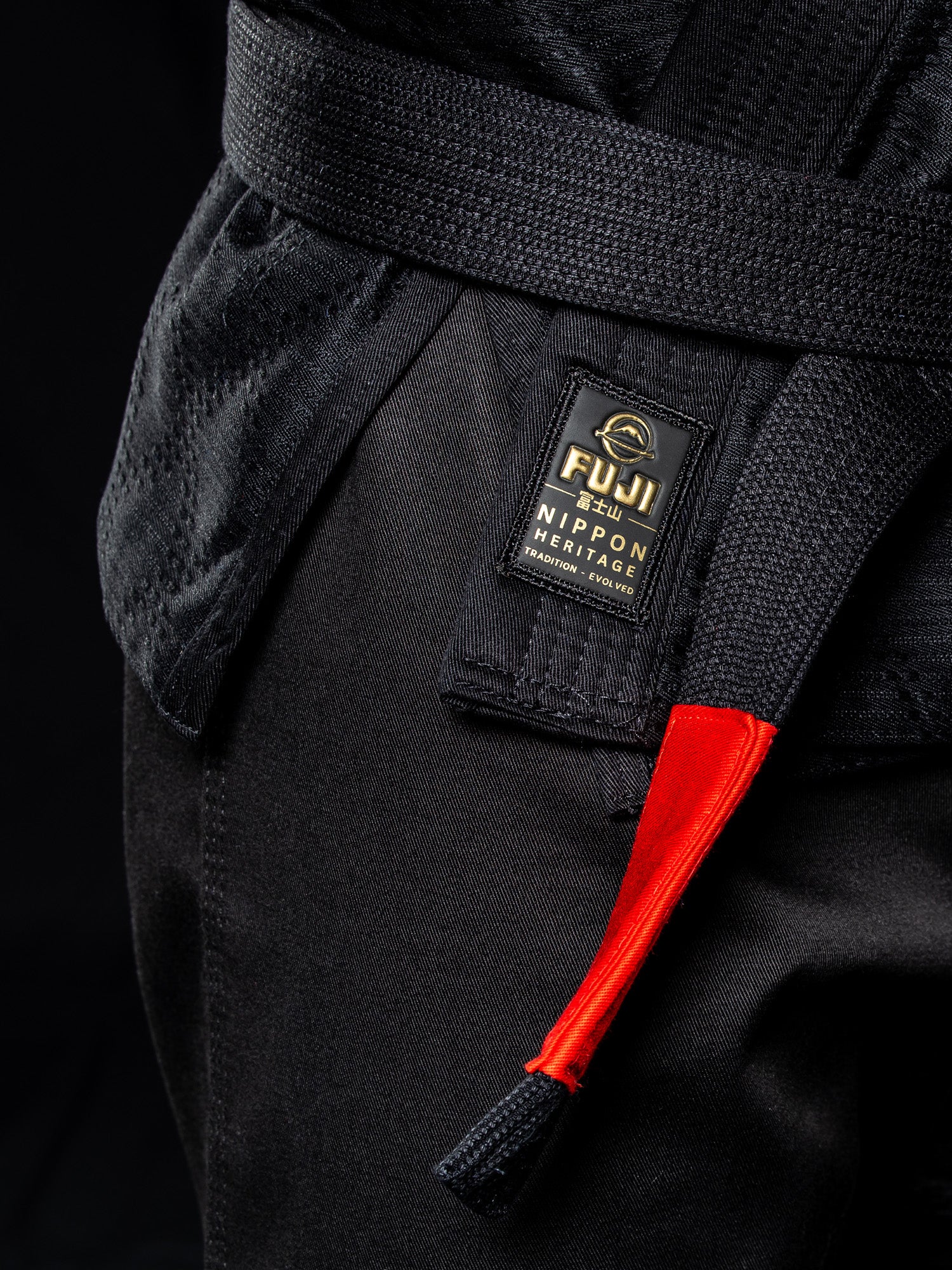 Close-up of the Heritage Keisho Lightweight BJJ Gi in black, featuring a Fuji Nippon Heritage patch and a black belt with a red tag, set against a dark background.