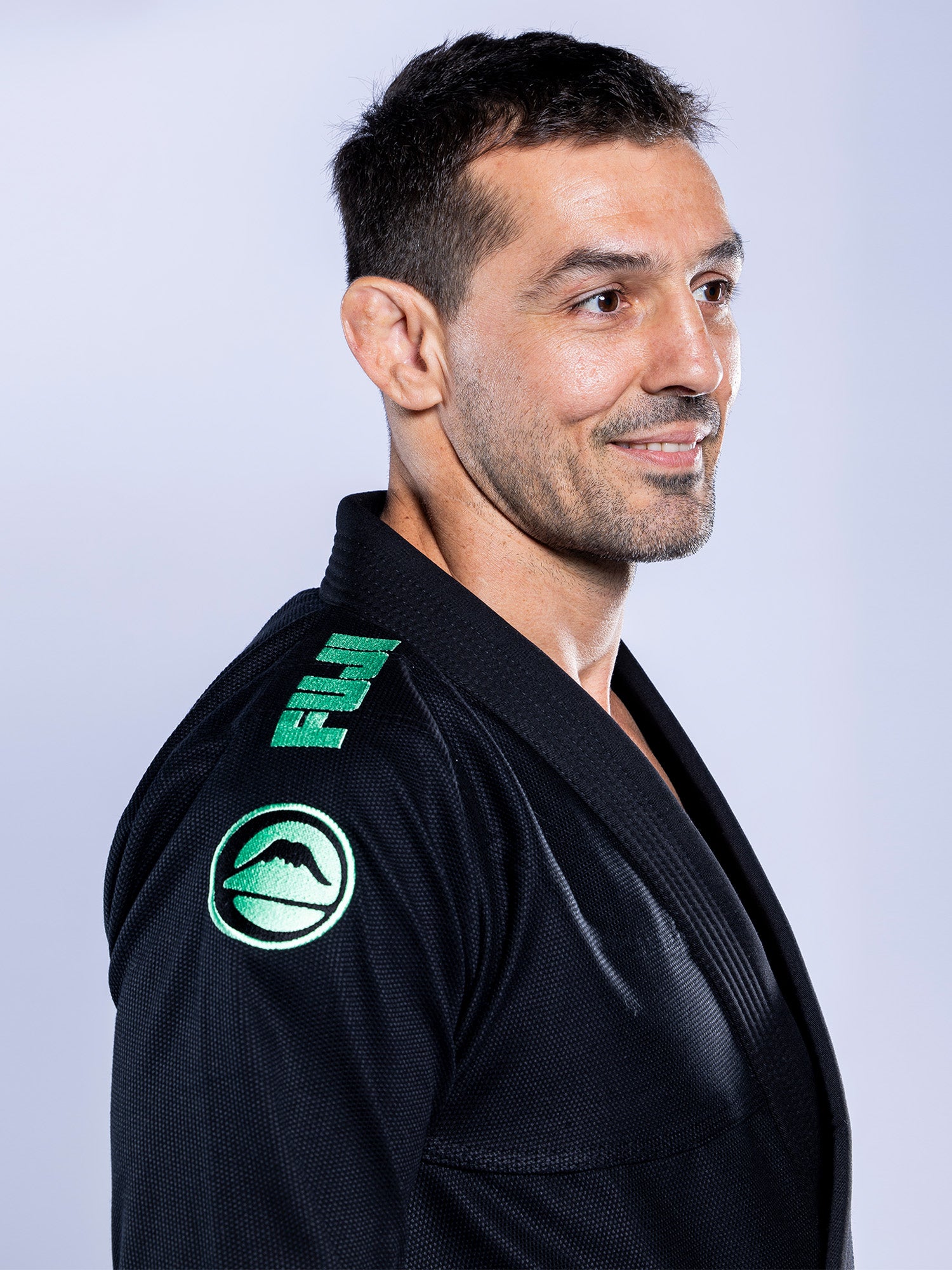A man wears the Baseline 2.0 BJJ Gi Black Mint, featuring green mint patches on the shoulder and sleeve, standing in profile and smiling against a light, plain background.