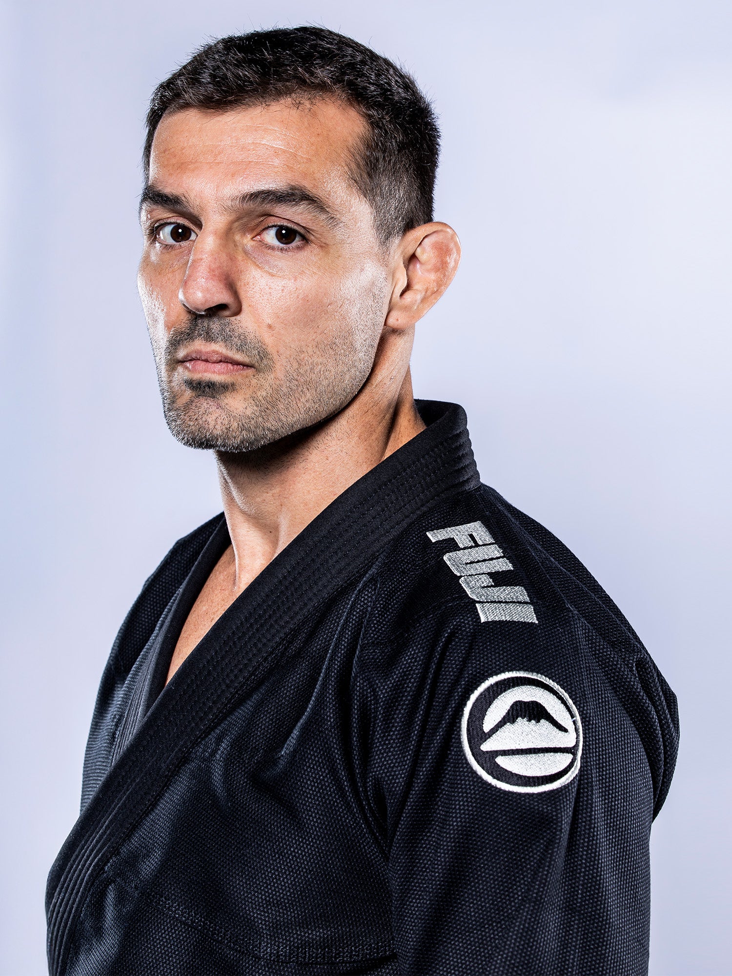 A man wears the Baseline 2.0 BJJ Gi Black Grey with a FUJI mountain logo on the shoulder, looking seriously at the camera against a plain light background.