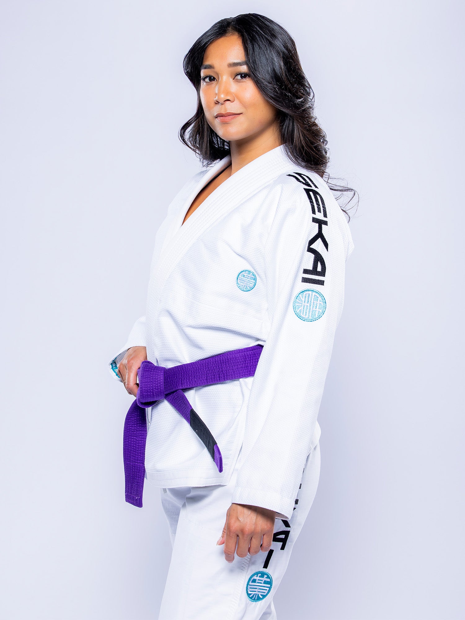 A woman with long dark hair stands confidently against a plain light background, wearing the Womens SEKAI 3.0 BJJ Gi in white and teal with a purple belt.