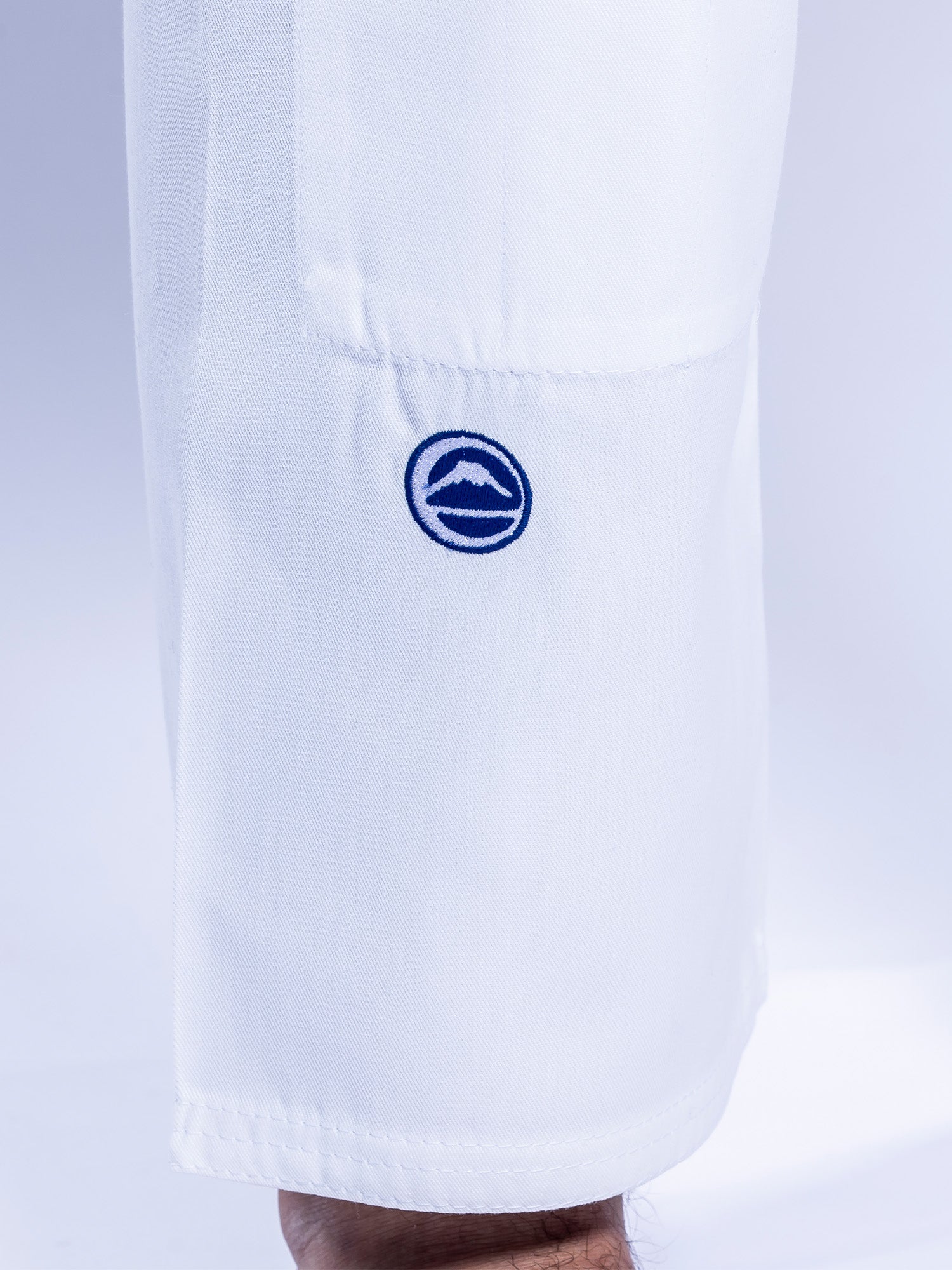 A close-up shows the lower part of the Baseline 2.0 BJJ Gi White Navy pant leg with a blue circular mountain logo embroidered near the hem; part of the wearer's foot appears at the bottom.