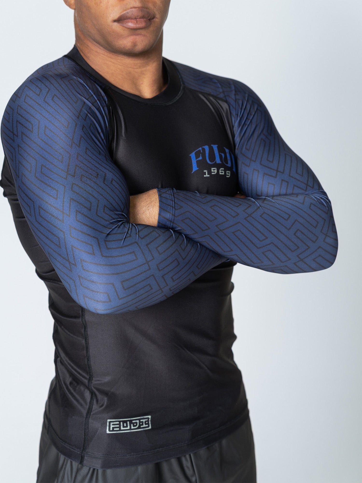 A person wearing the Oni Samurai Flex Lite Rashguard Blue, featuring black and blue colors with geometric patterned sleeves and "FUDI 1969" on the chest, stands with arms crossed; their face is partially out of frame.