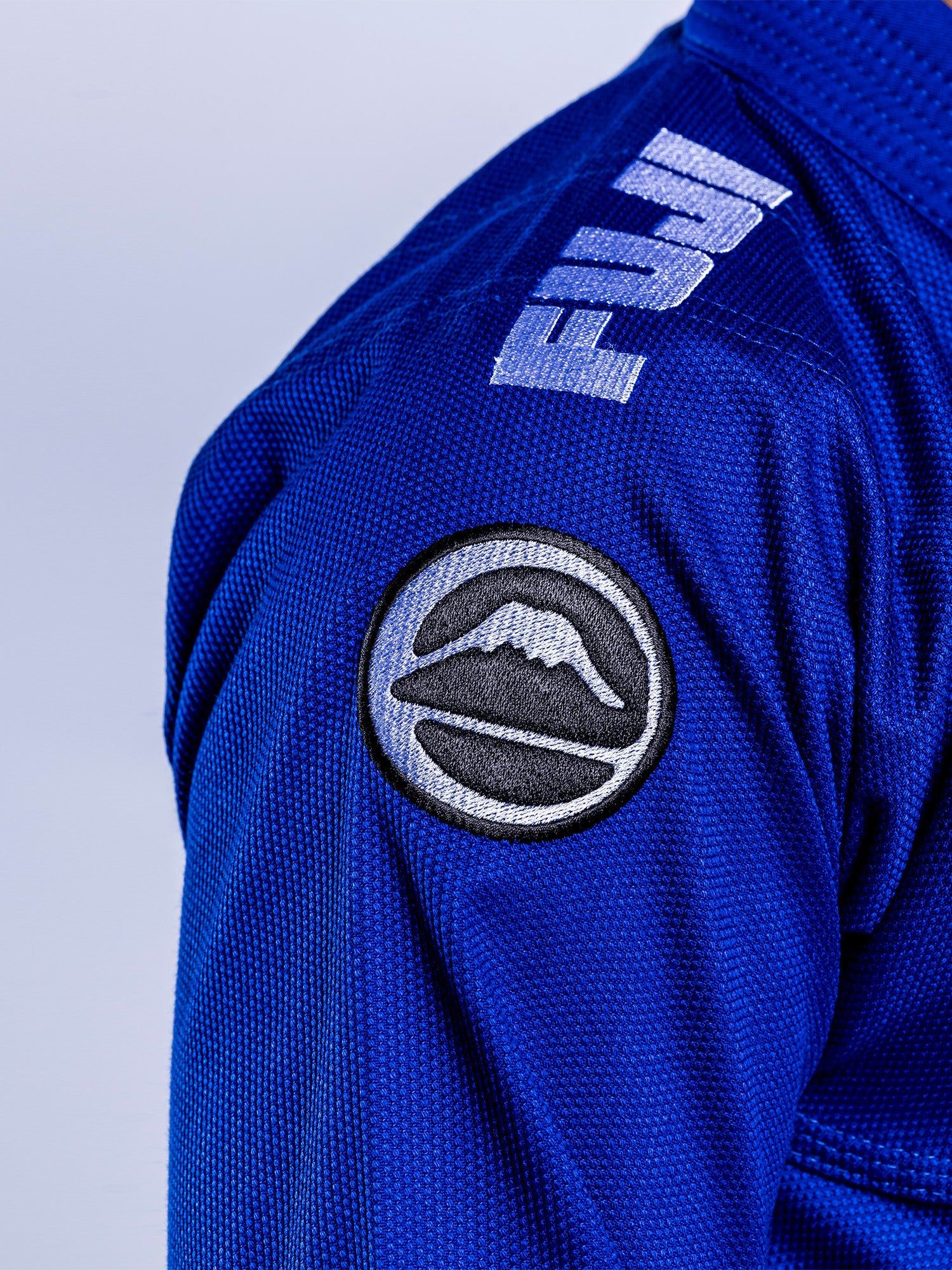 Close-up of a person wearing the Baseline 2.0 BJJ Gi Blue White with "FUJI" embroidered on the shoulder and a circular mountain logo patch on the upper arm.