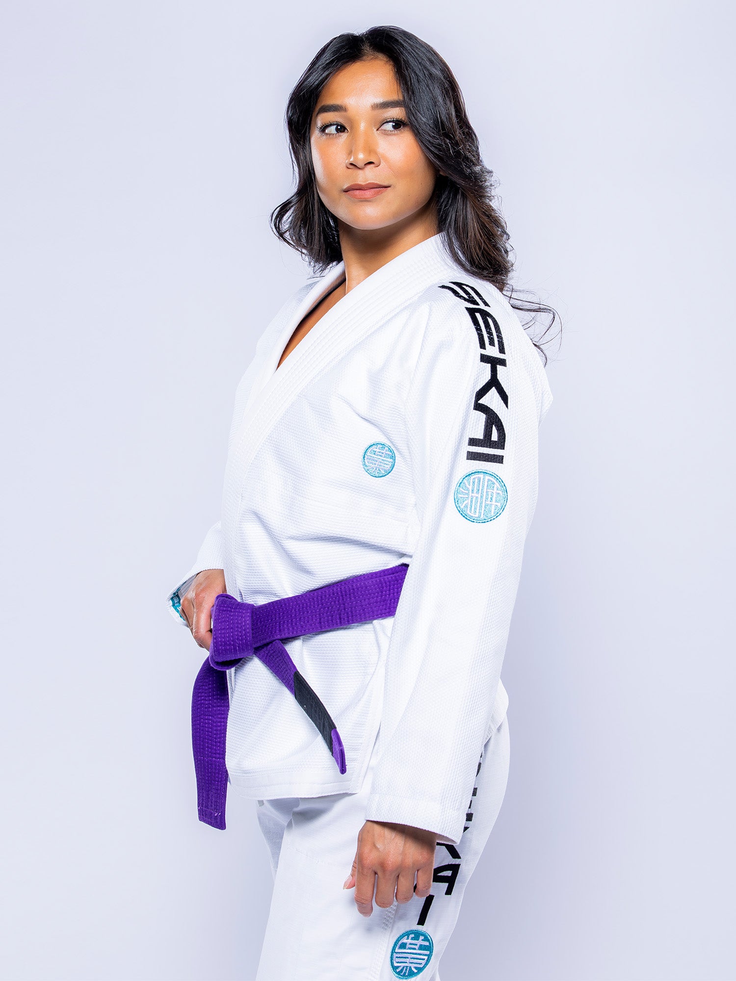 A woman with long, dark hair wears the Womens SEKAI 3.0 BJJ Gi in white and teal, paired with a purple belt, standing in profile against a light background and looking confidently to the side.