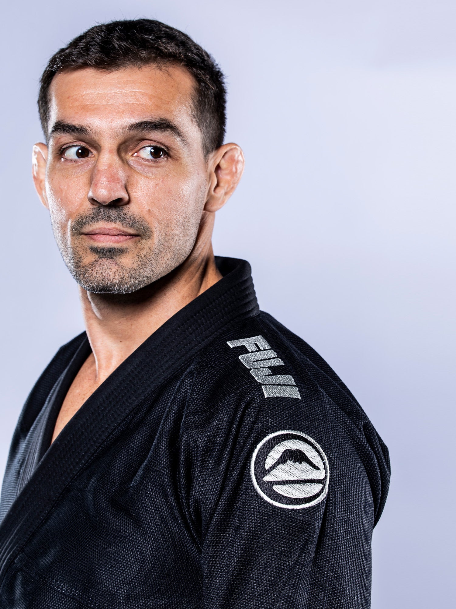 A man with short dark hair and stubble wears the Baseline 2.0 BJJ Gi Black Grey, featuring a Fuji logo on the shoulder, looking to his left against a light gray background.