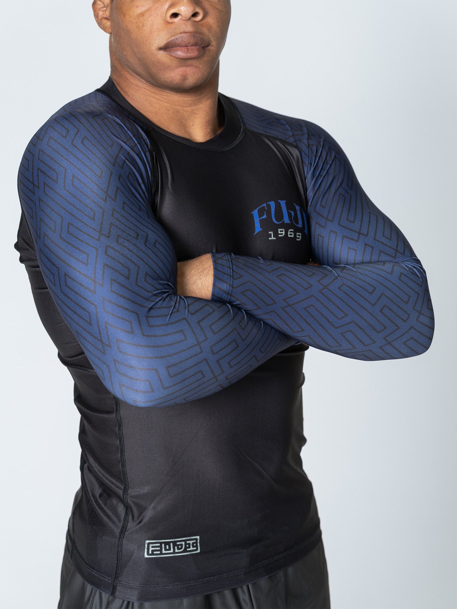 A person with crossed arms wearing the Oni Samurai Flex Lite Rashguard Blue, a fitted black and blue geometric-patterned long-sleeve athletic shirt with FWD 1969 text on the chest and a small logo at the waist.