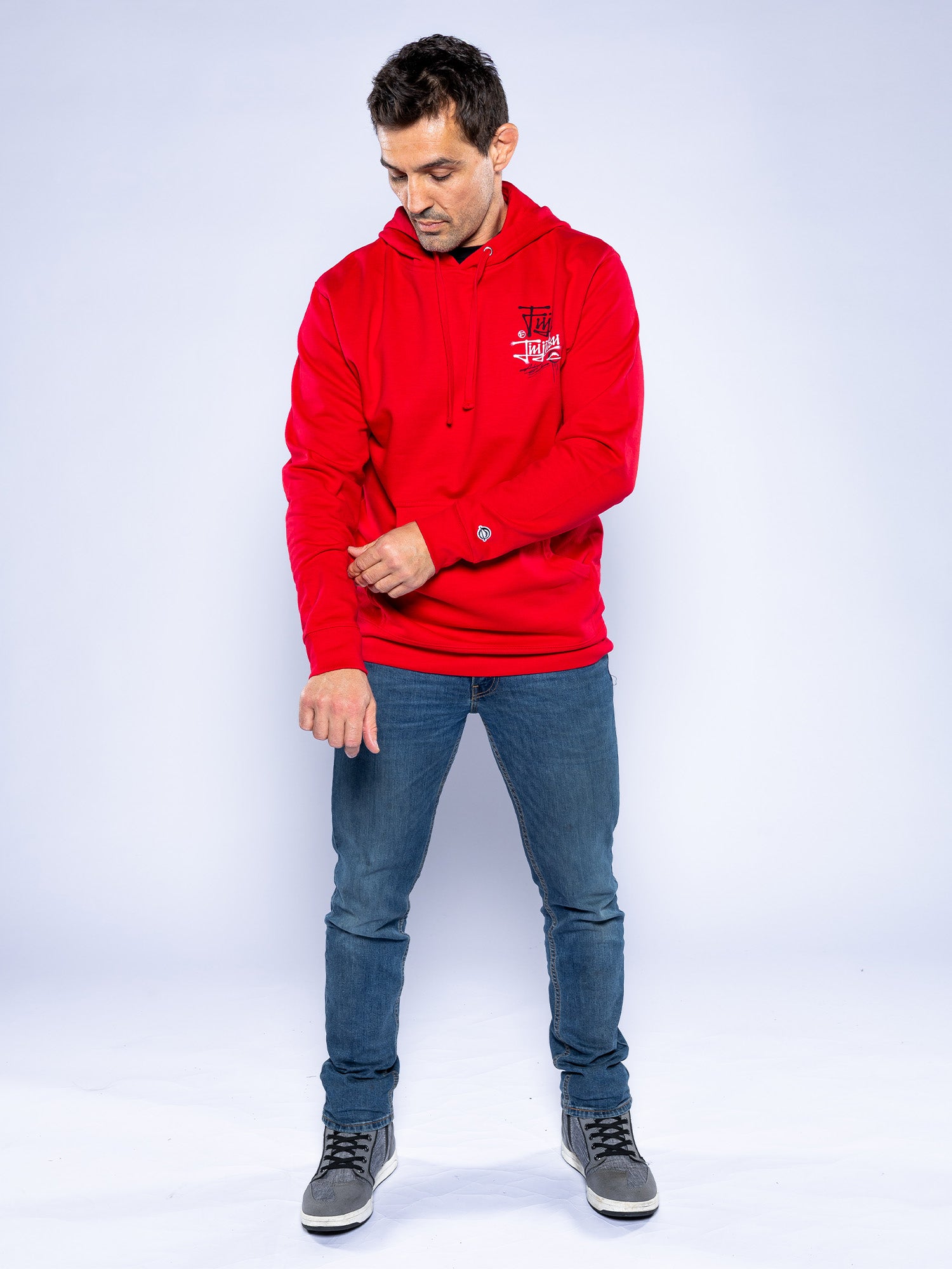 A man wearing the Japanese Street Art Hoodie Red, blue jeans, and gray sneakers stands against a plain white background, adjusting his left sleeve while looking down.