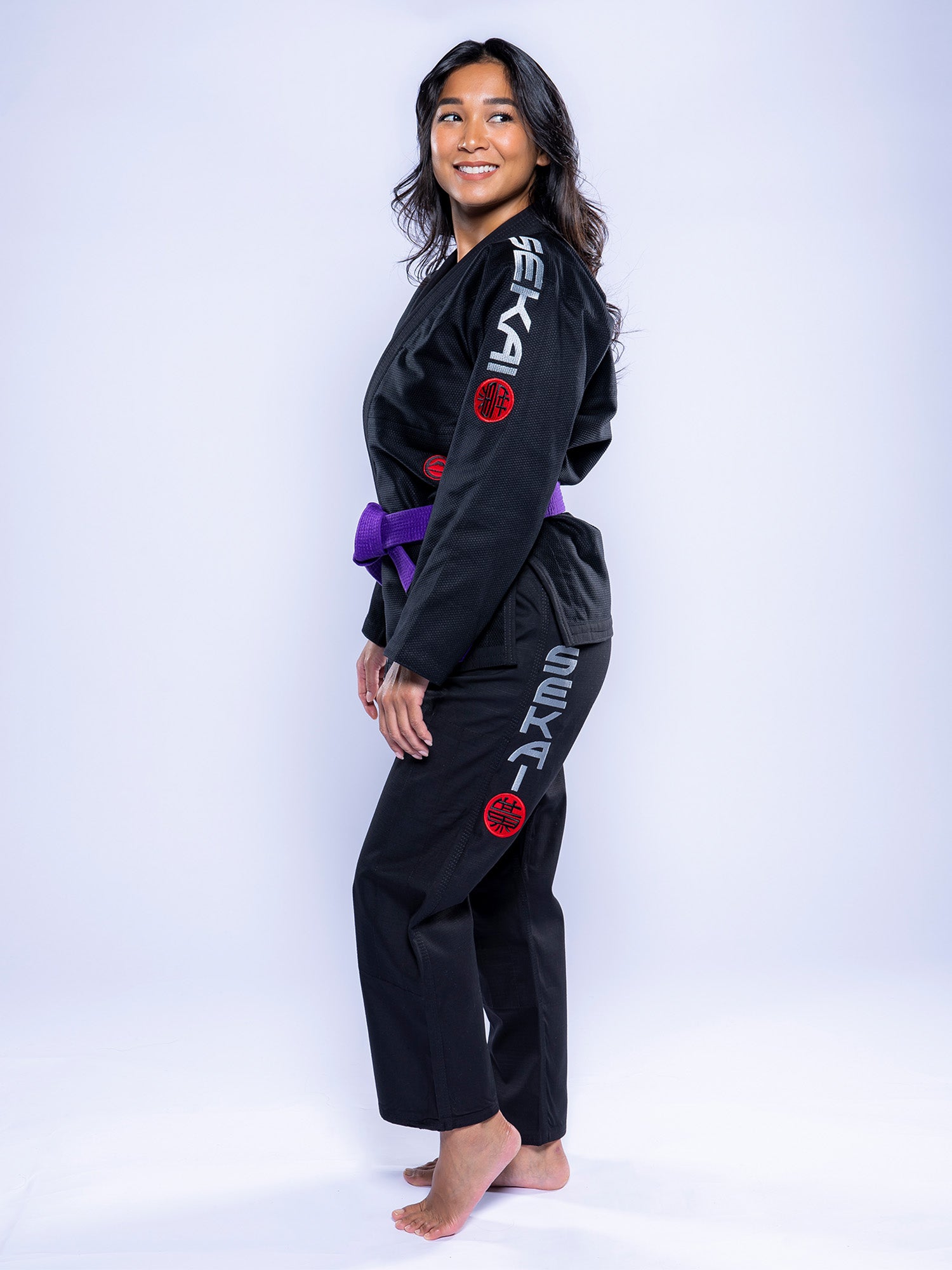 A woman wears the Womens SEKAI 3.0 BJJ Gi Black Grey, featuring SEKAI on the sleeve and pant leg, a purple belt, and stands barefoot smiling against a white background.