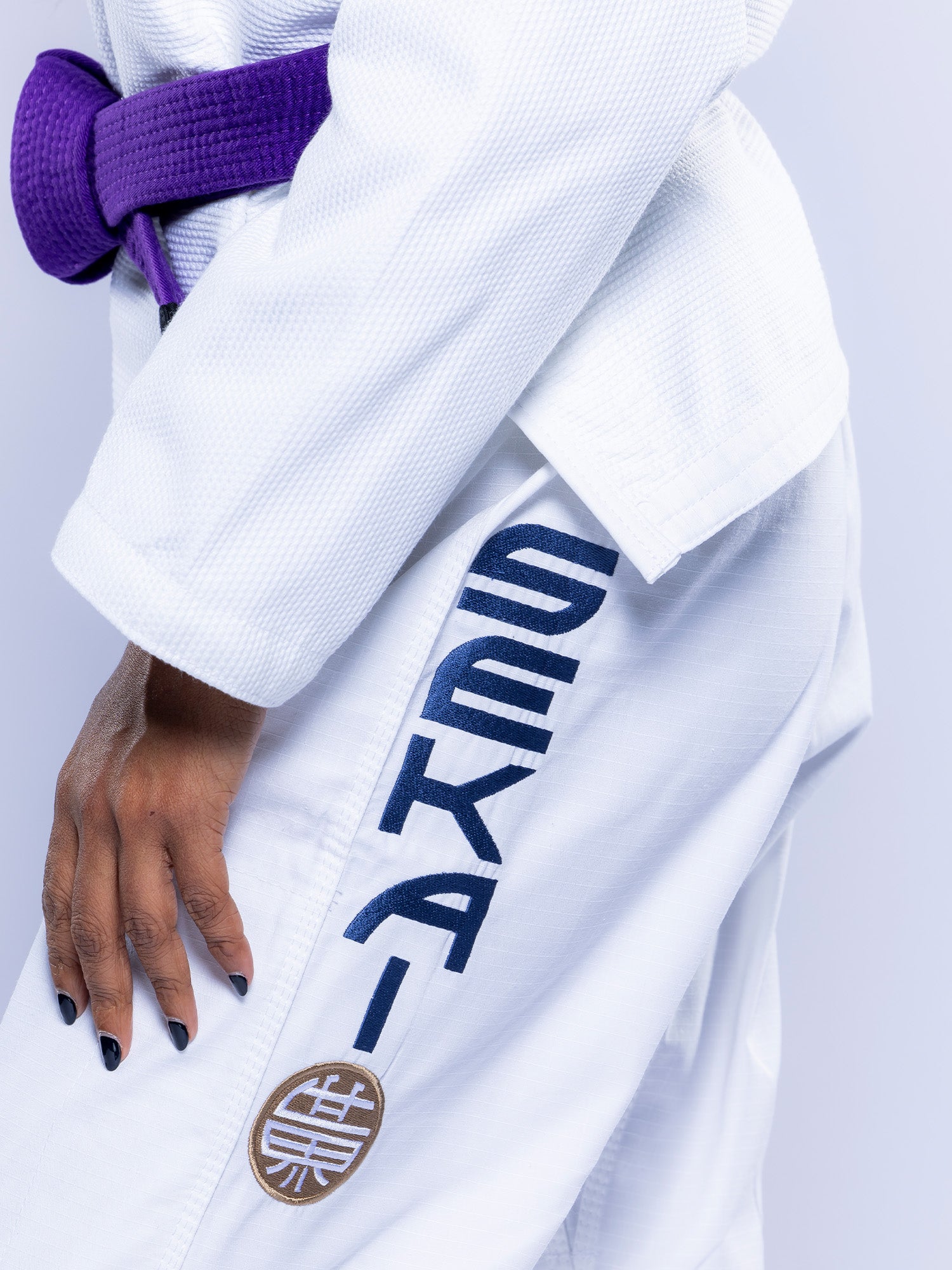 A person wears the Womens SEKAI 3.0 BJJ Gi White Navy, featuring "SEKAI" in bold and a circular patch with Japanese characters on the pants, paired with a purple belt.
