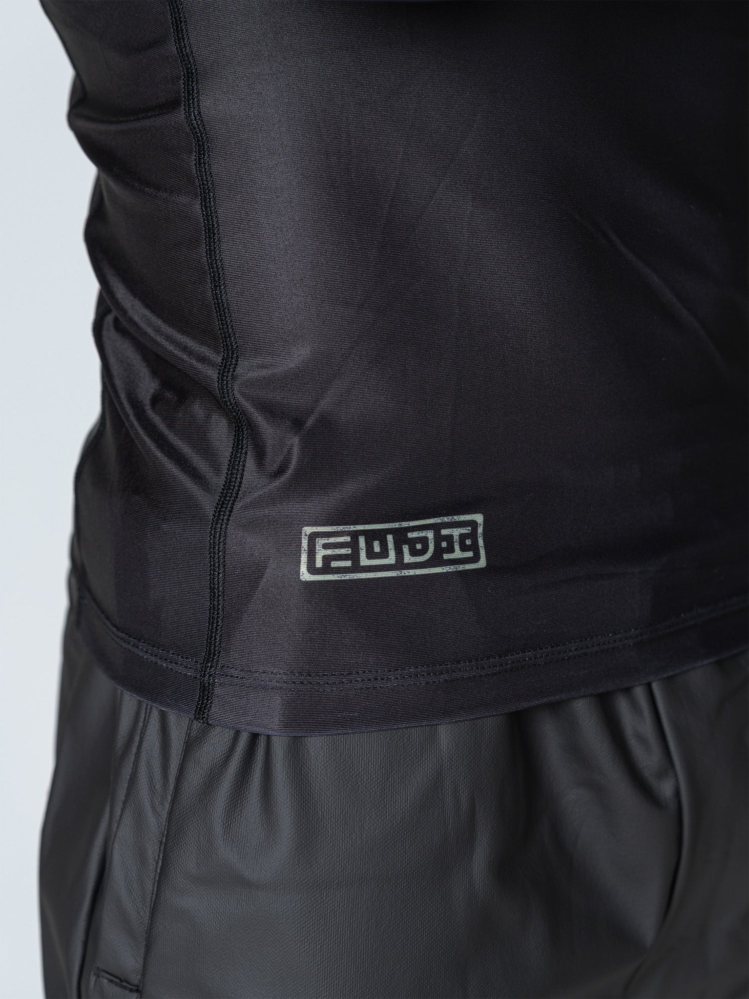 A close-up of a person wearing the Oni Samurai Flex Lite Rashguard Blue, a shiny black and blue athletic shirt with a small rectangular FUBU logo label stitched near the hem.