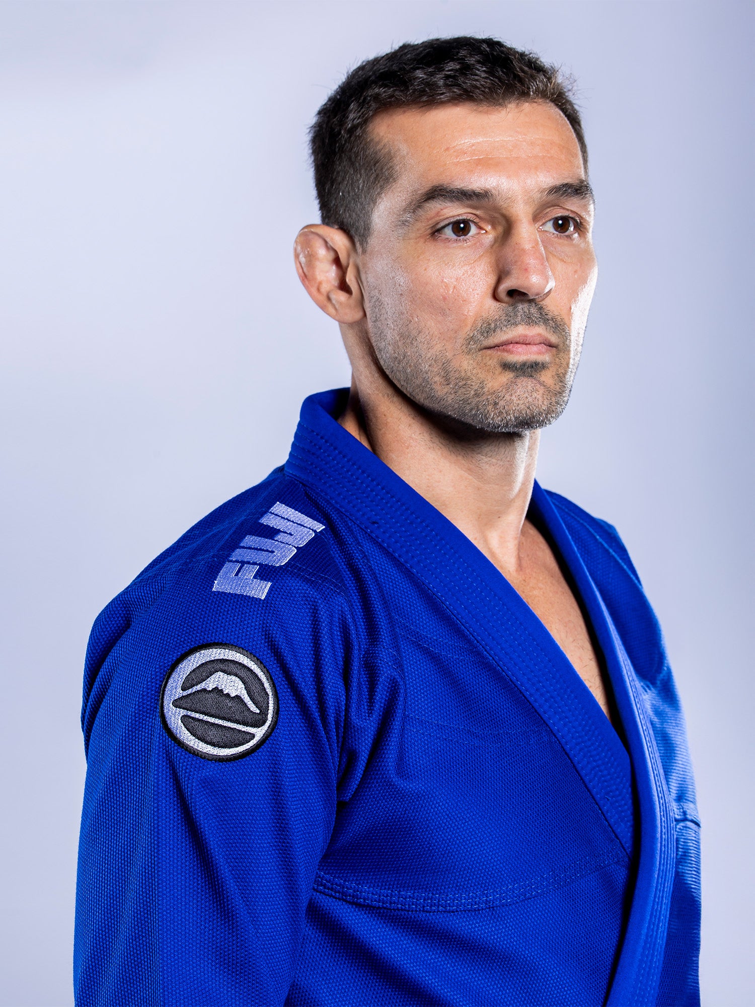 A man in a blue Baseline 2.0 BJJ Gi with white accents and a Fuji logo stands against a plain light background, looking serious and focused.