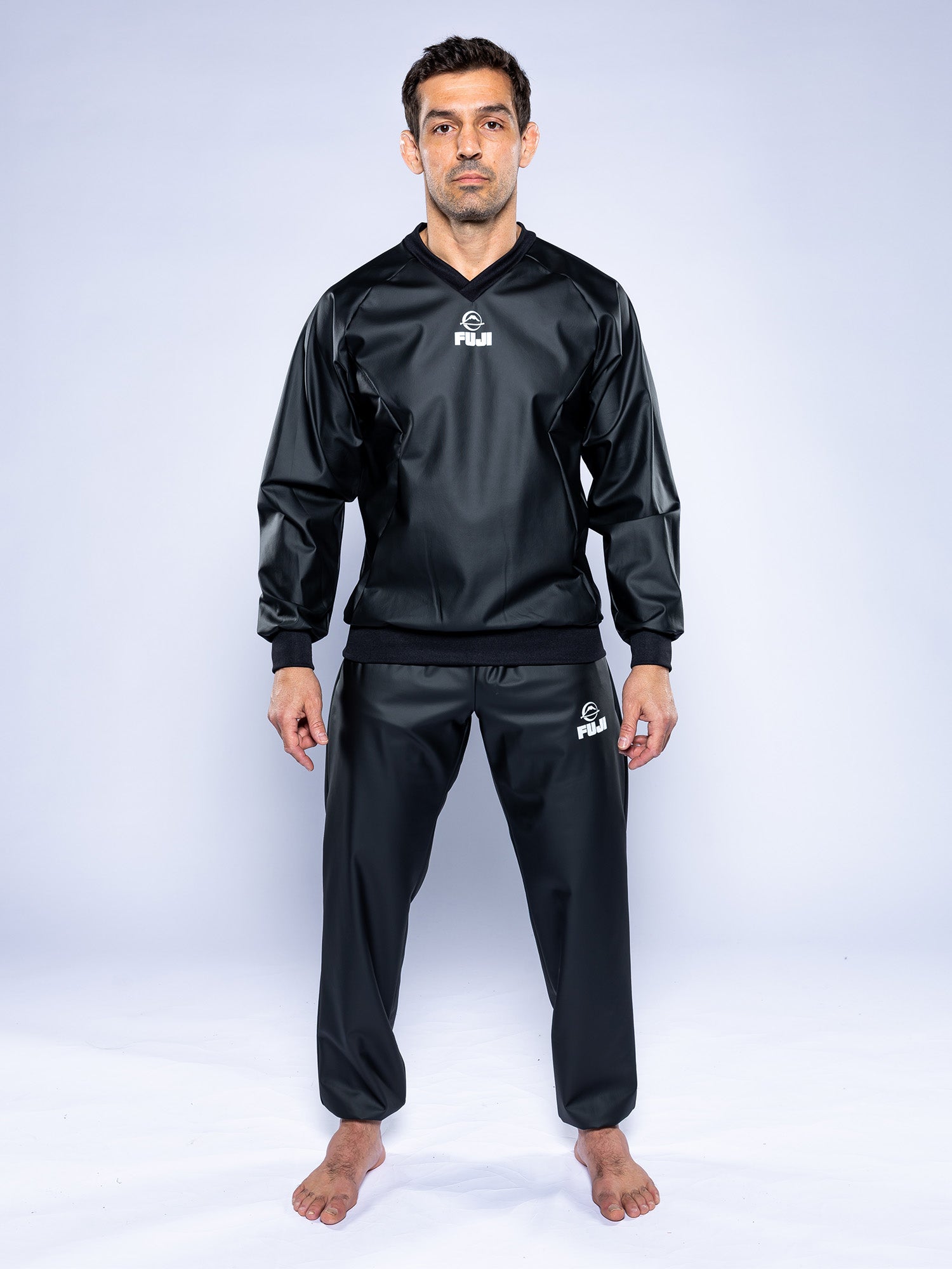 A man stands barefoot on a white background, wearing a black Grappling Sauna Suit with the logo on the chest and left thigh. He faces forward with a neutral expression, arms relaxed at his sides.