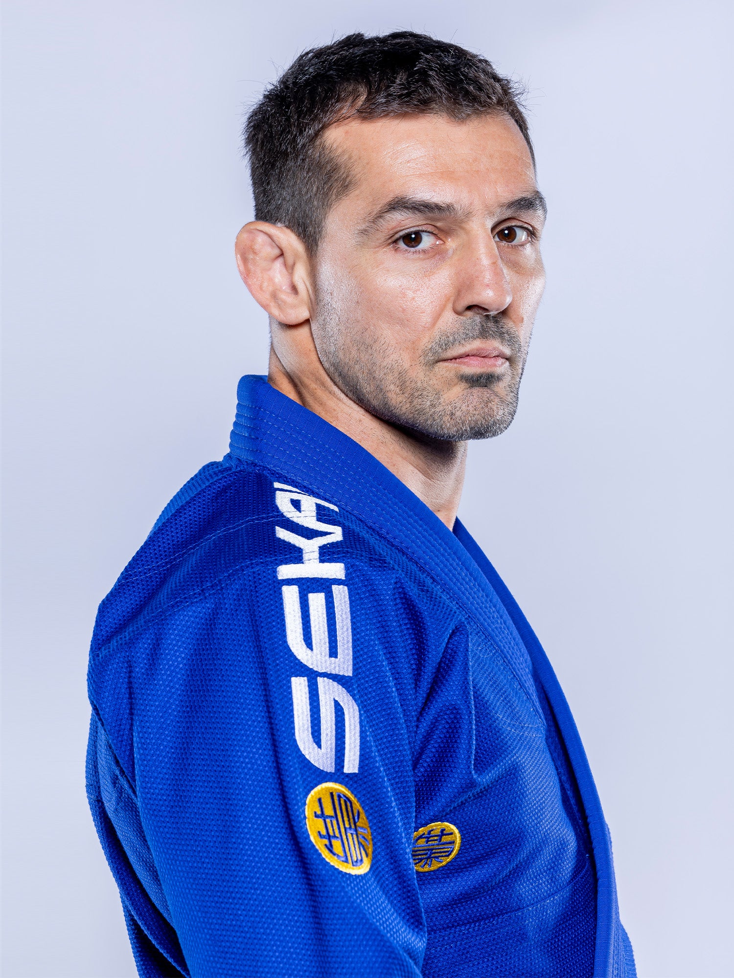 A man with short dark hair and stubble wears the SEKAI 3.0 BJJ Gi Blue Yellow, featuring white SEKA lettering on the shoulder and yellow patches, looking seriously at the camera against a plain light background.