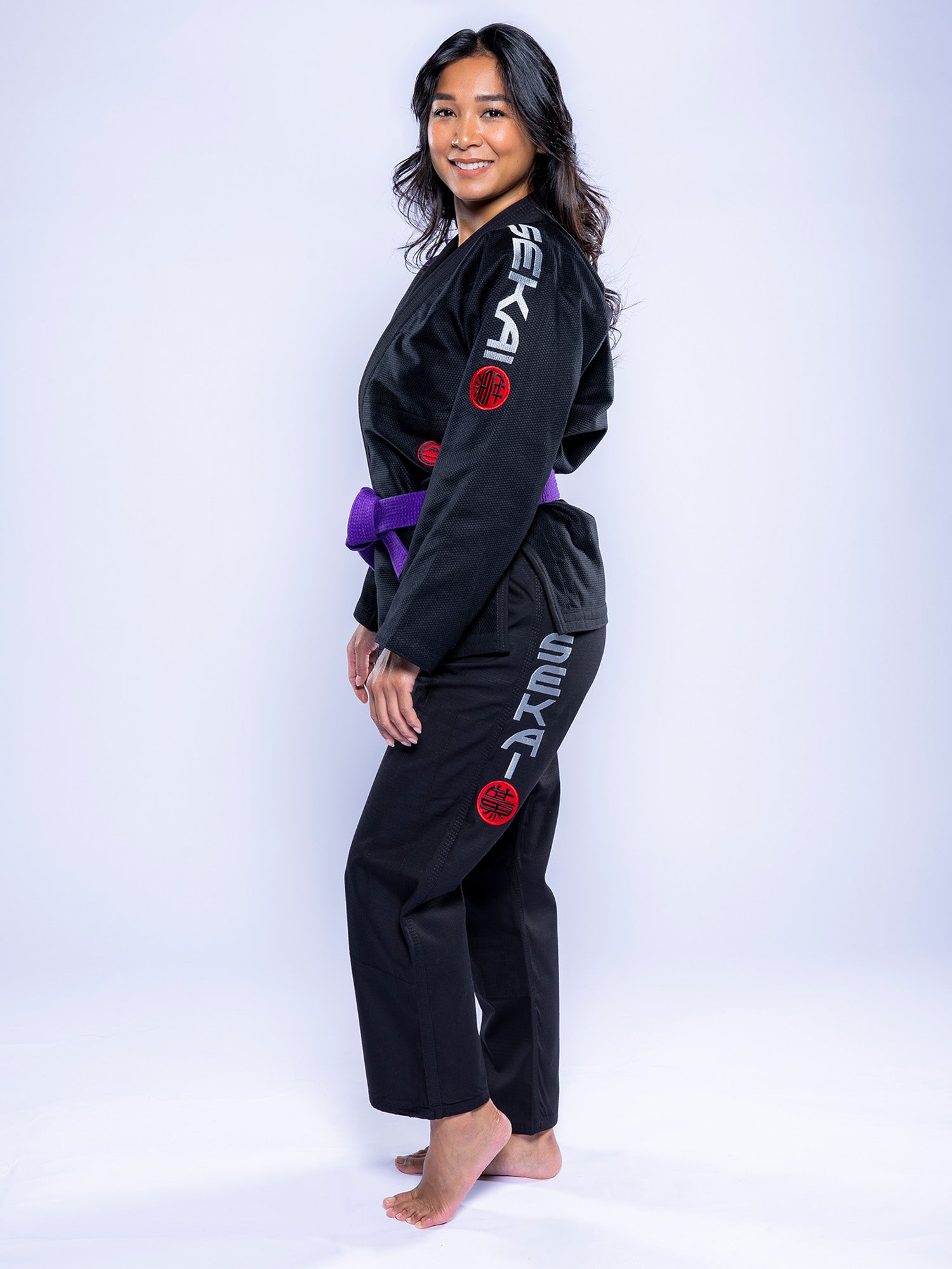 A woman wearing the Womens SEKAI 3.0 BJJ Gi Black Grey with a purple belt stands barefoot, smiling and posing against a plain white background. The gi features gray SEKA lettering and red circular patches on the sleeve and pants.