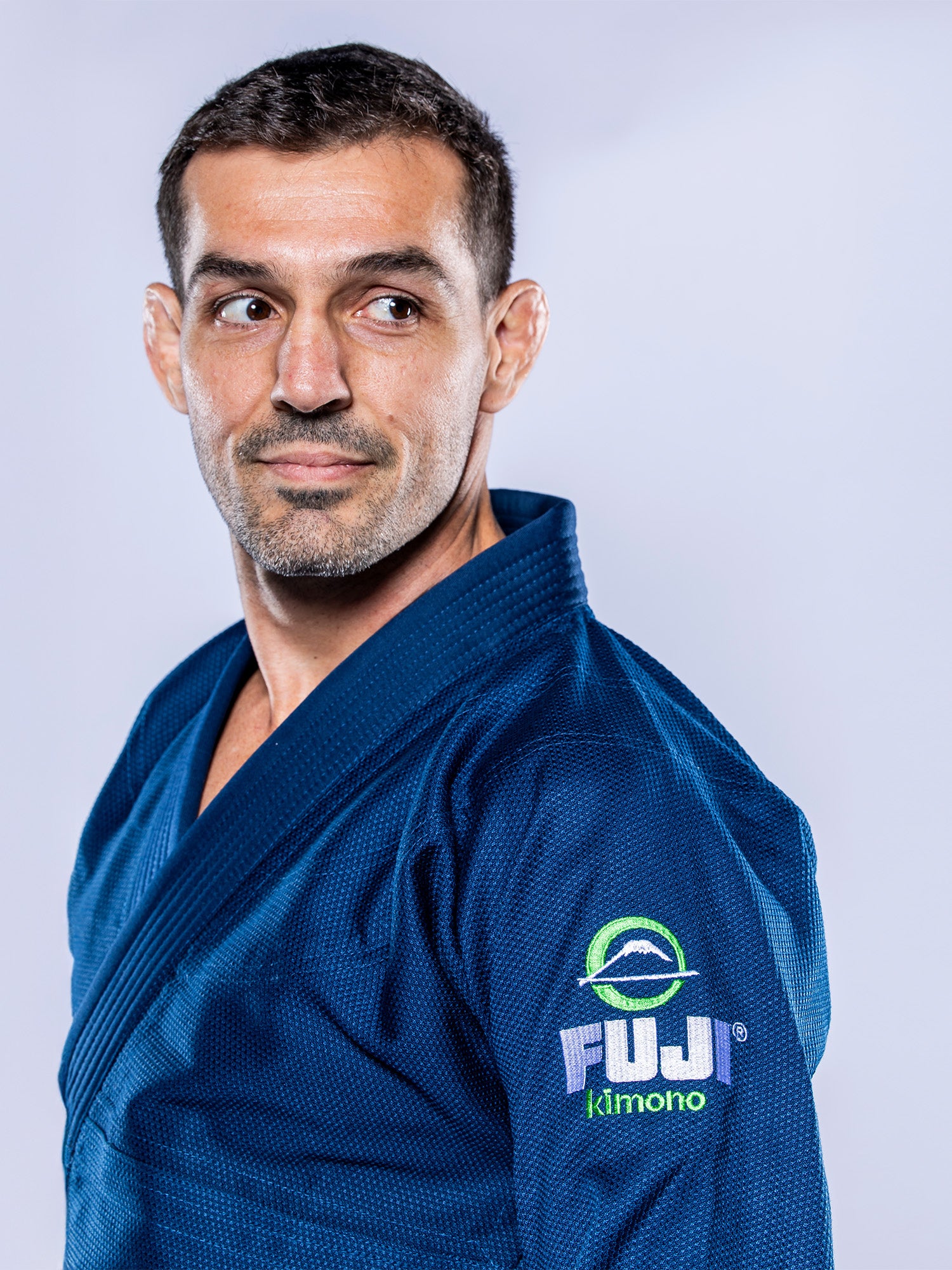 A man with short dark hair and a trimmed beard wears the Lightweight BJJ Gi Navy, featuring a Fuji Kimono logo on the sleeve, and looks to his left against a light grey background.