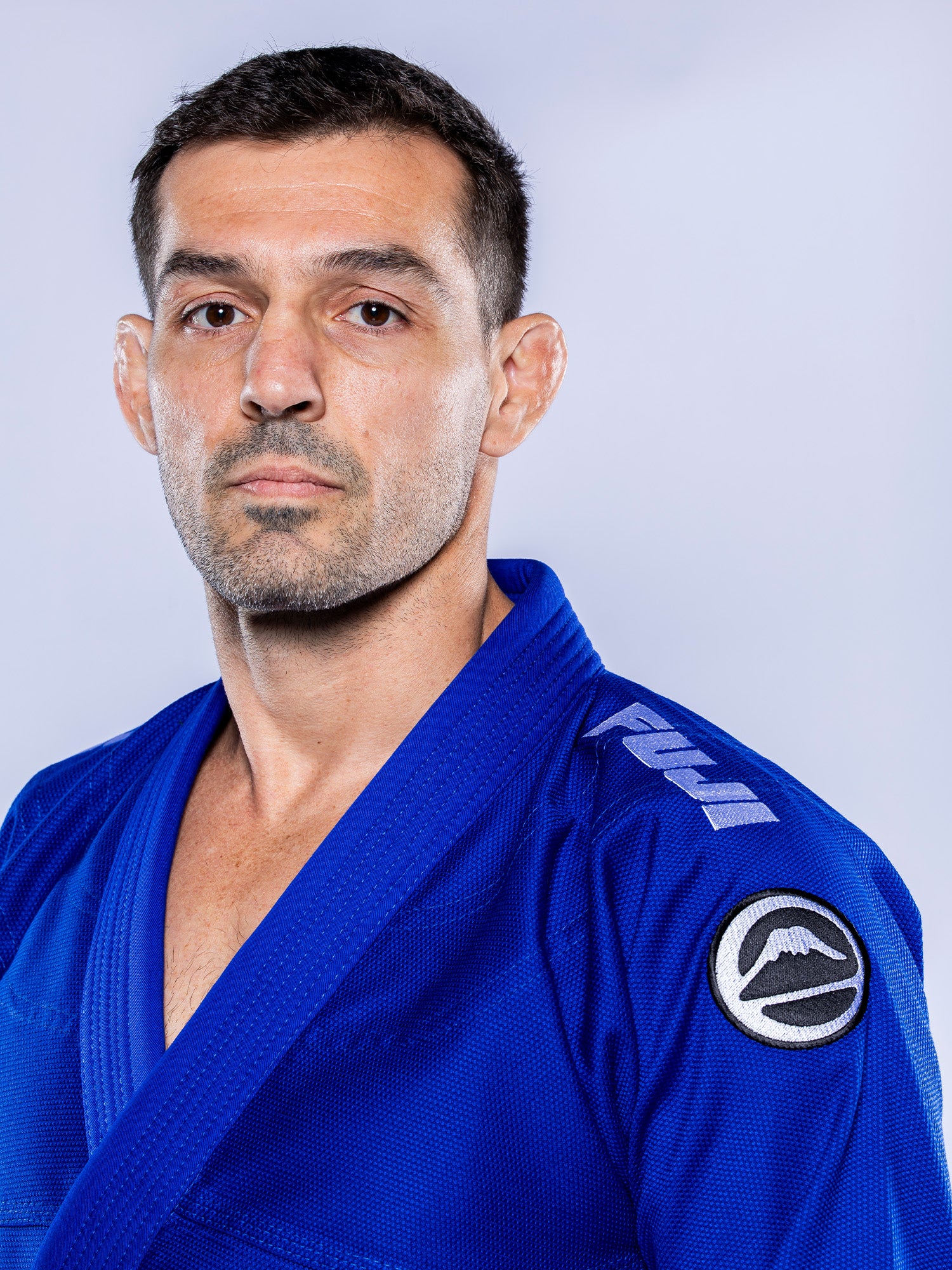 A man with short dark hair wears the Baseline 2.0 BJJ Gi Blue White, featuring a Fuji logo on the shoulder, looking serious and facing slightly left against a plain light background.