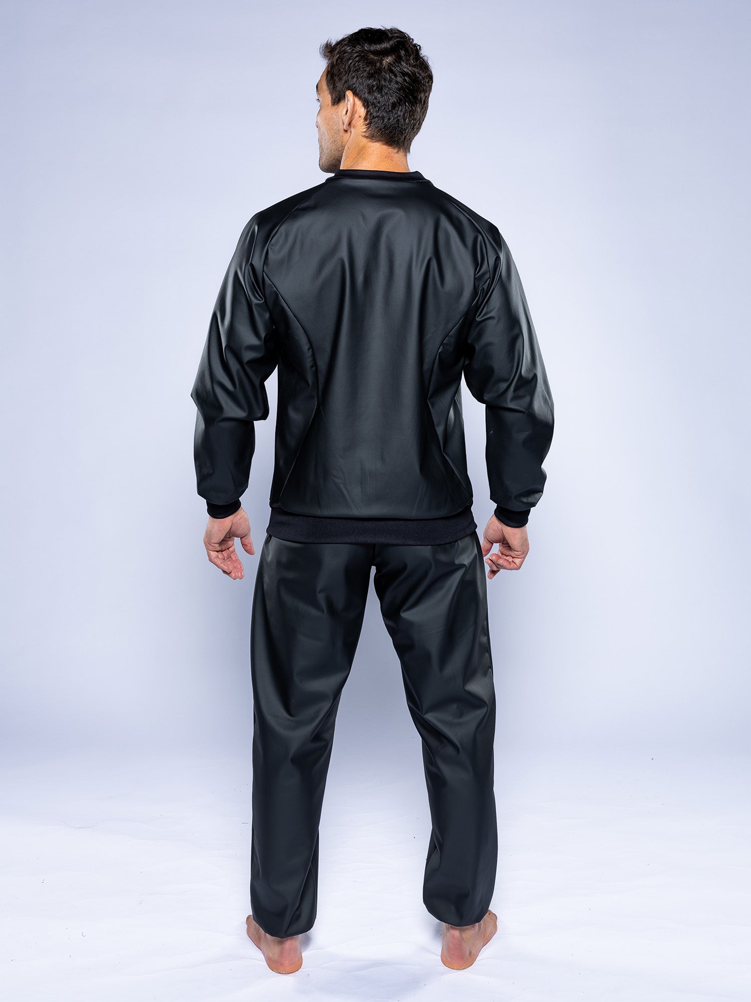 A person stands barefoot on a white background, facing away from the camera, wearing the Grappling Sauna Suit Black with long sleeves and pants.