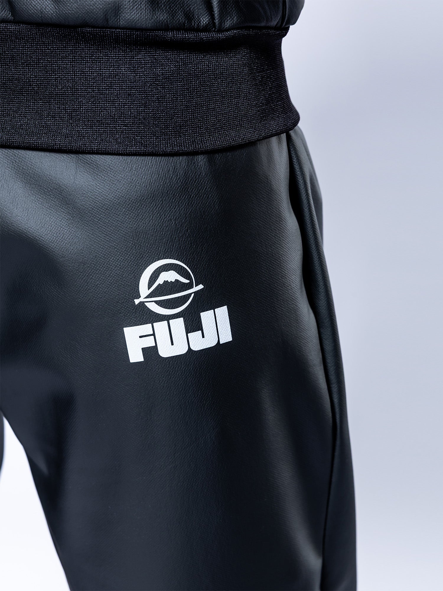 Close-up of a person wearing the Grappling Sauna Suit Black, featuring a white FUJI logo and mountain graphic on the upper thigh. The background is plain and light-colored.