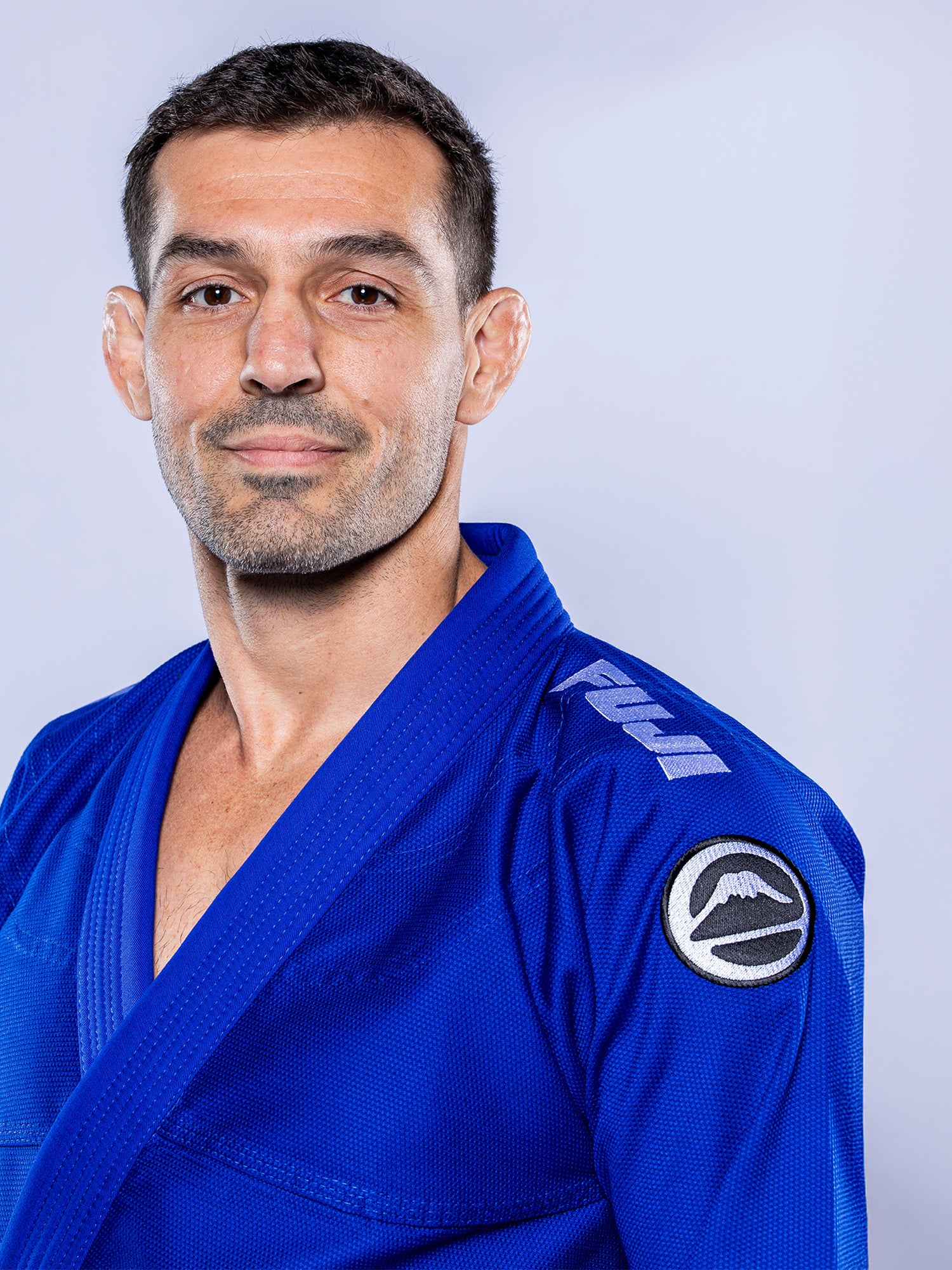 A man with short dark hair and light stubble models the Baseline 2.0 BJJ Gi Blue White, which features shoulder and sleeve patches, against a plain light gray background.