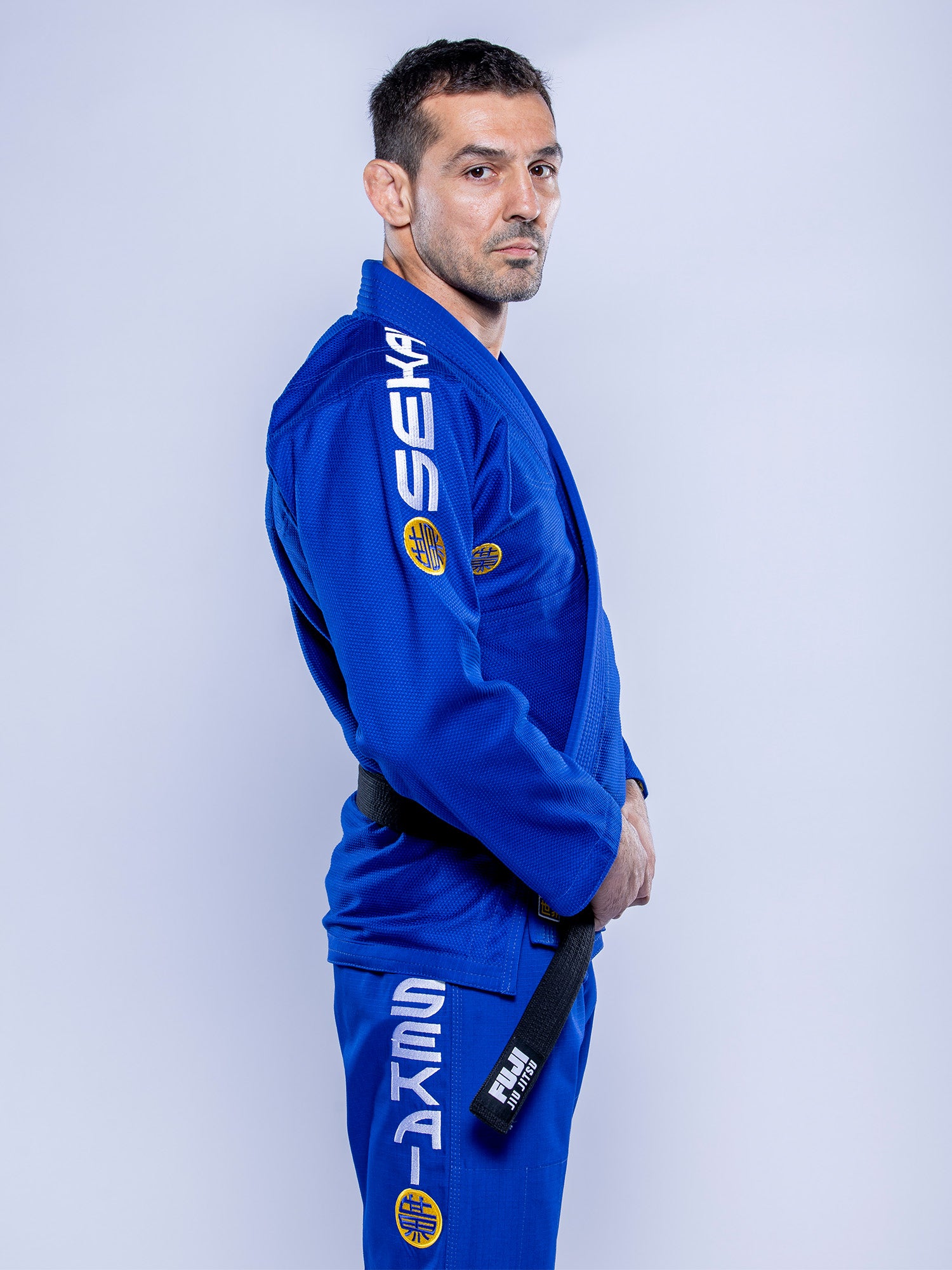 A man wearing the SEKAI 3.0 BJJ Gi Blue Yellow stands in profile, holding his black belt and looking seriously at the camera against a plain light background.
