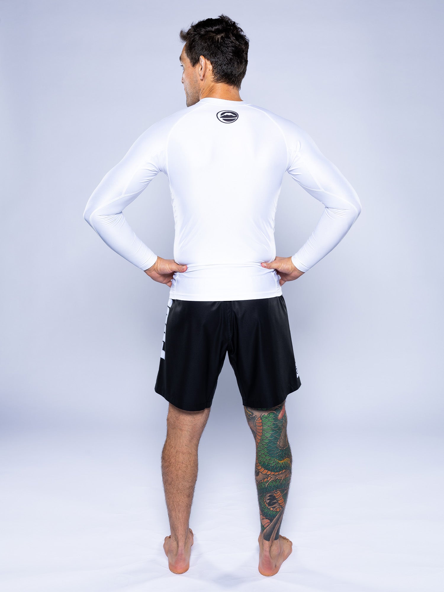 A man stands barefoot with his back to the camera, wearing the Baseline 2.0 Long Sleeve Rashguard in white and black shorts. His right leg features a vibrant, colorful tattoo design.