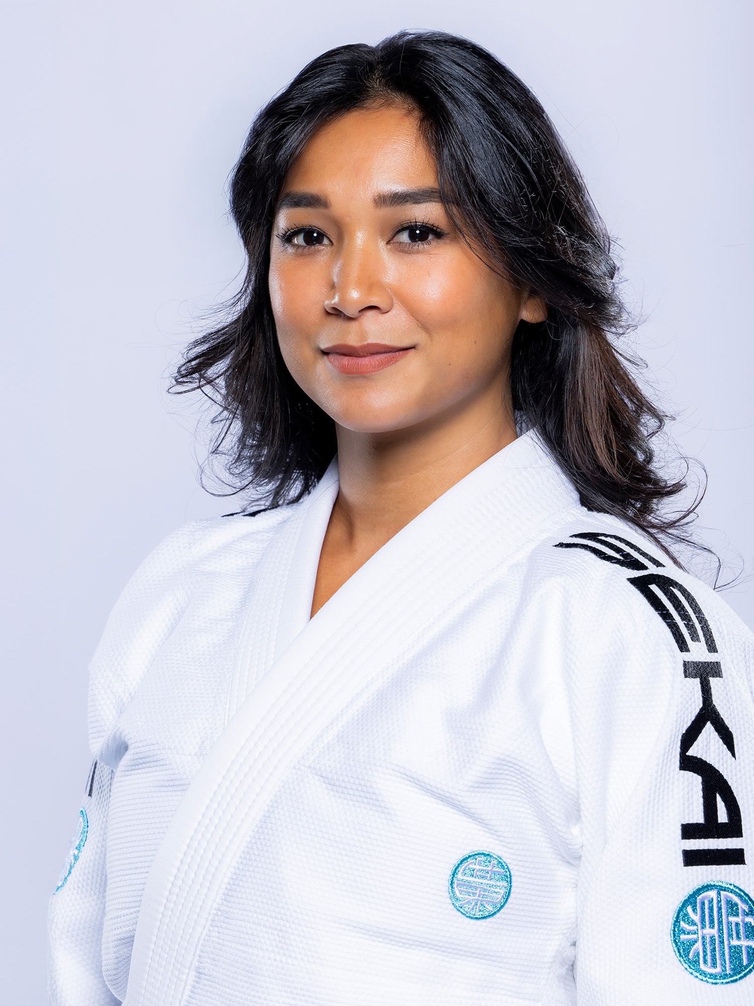 A woman with long, dark hair smiles softly at the camera, wearing the Womens SEKAI 3.0 BJJ Gi White Teal against a plain light background.