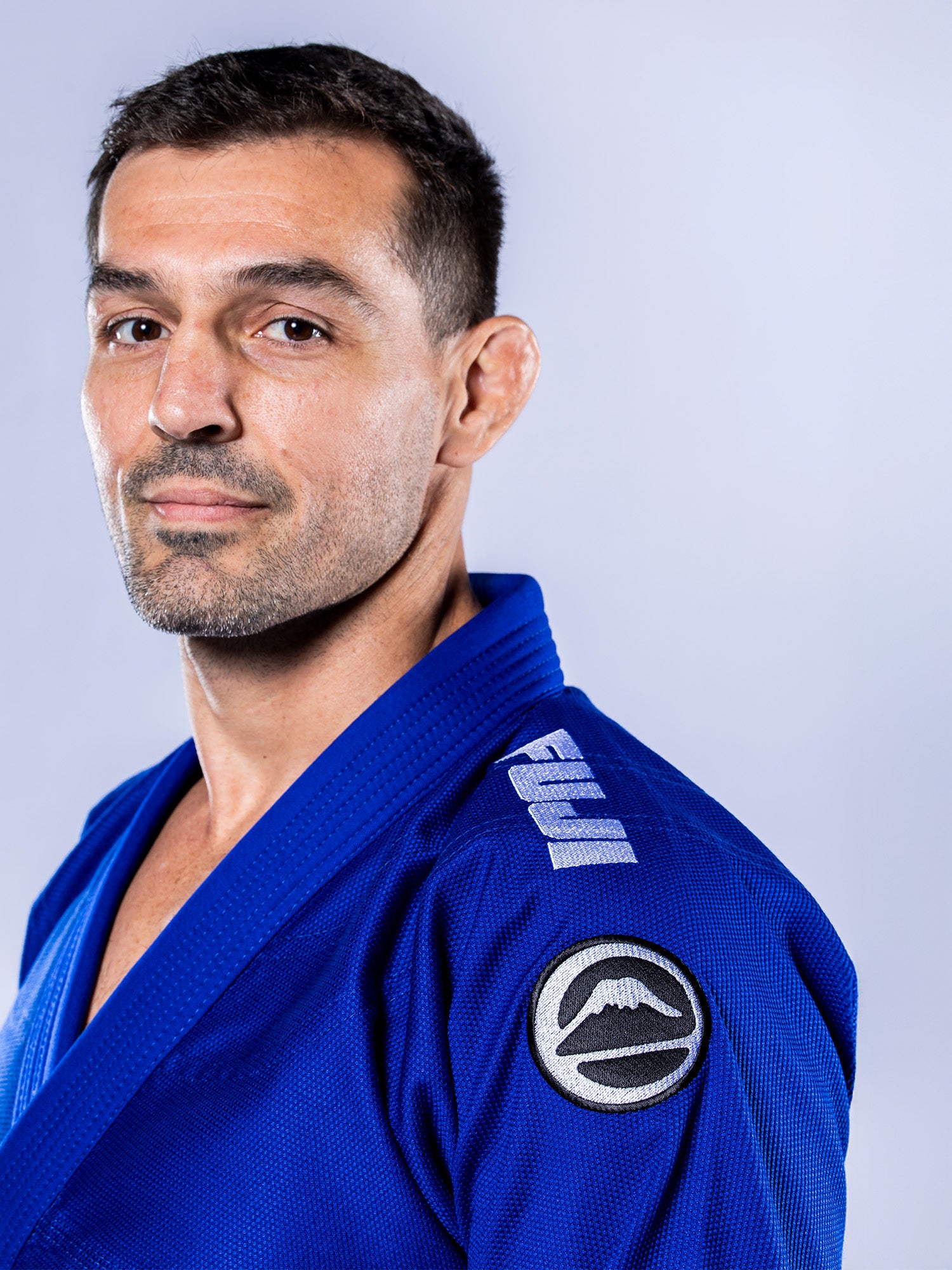 A confident man with short dark hair and a trimmed beard wears the Baseline 2.0 BJJ Gi Blue White, featuring shoulder patches, and poses against a plain light background.