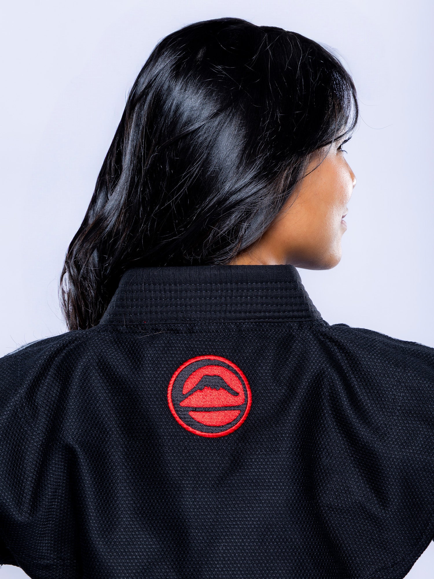 A woman with long dark hair is shown from behind, wearing the Womens SEKAI 3.0 BJJ Gi Black Grey, which features a red circular patch with a mouth and mustache on the back of the collar.