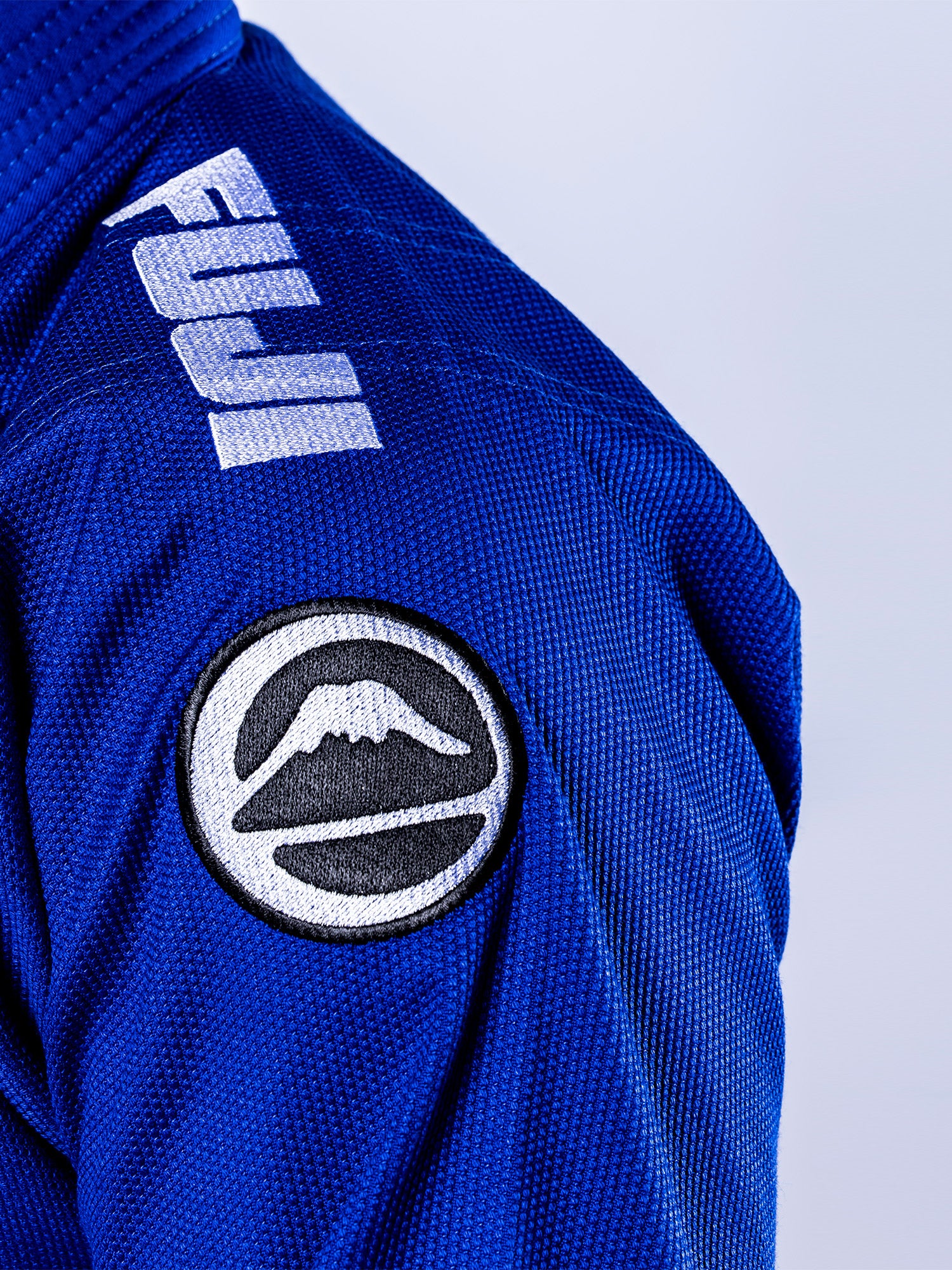 Close-up of the Baseline 2.0 BJJ Gi Blue White jacket, showing "FUJI" on the shoulder and a circular mountain logo patch on the upper arm.