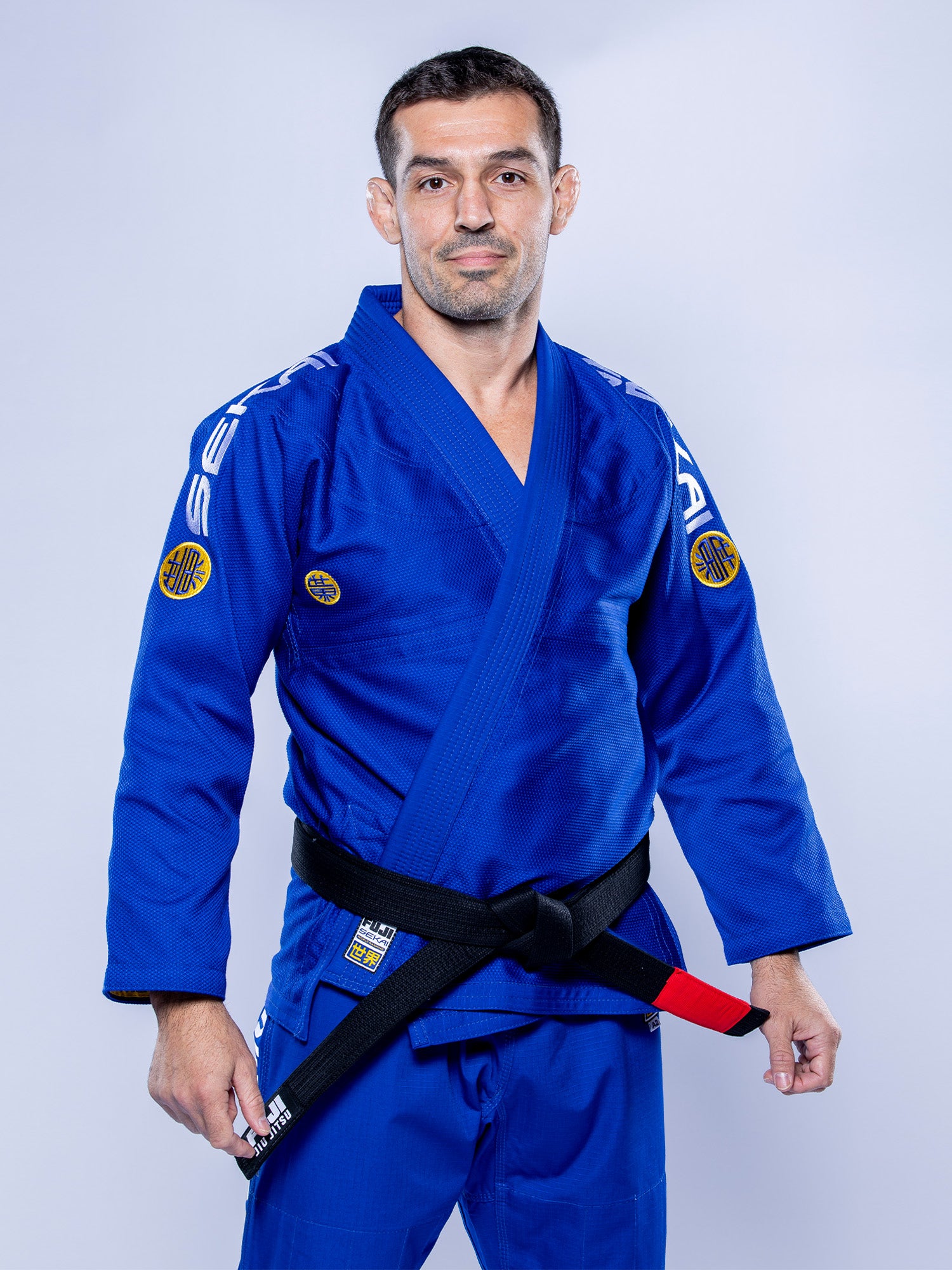 A man in the SEKAI 3.0 BJJ Gi Blue Yellow with a black belt stands confidently against a light grey background.