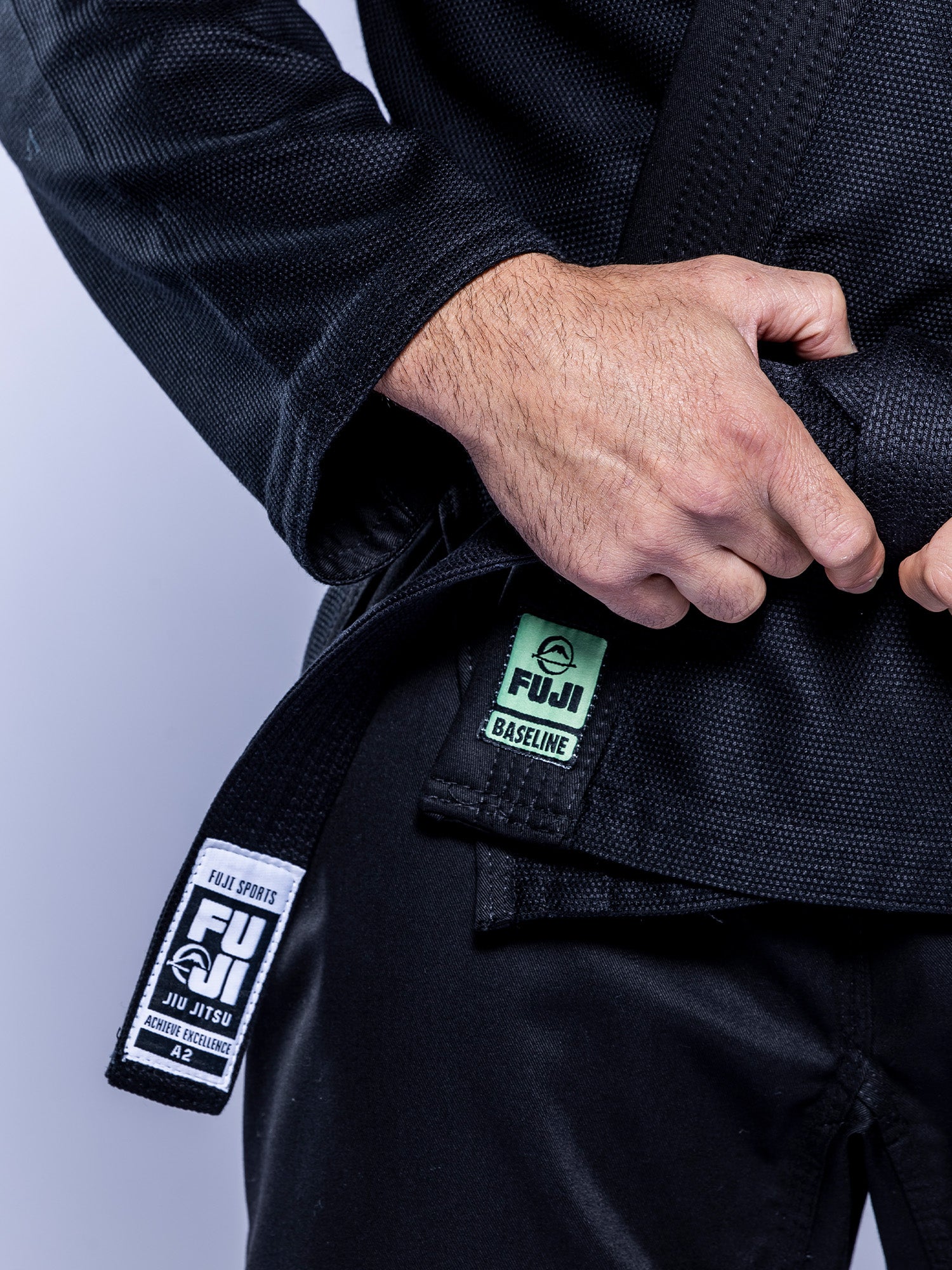 A person wearing the Baseline 2.0 BJJ Gi Black Mint adjusts their black belt. Fuji Sports and Baseline patches are visible near the waist; only the person's torso and hands can be seen in the image.