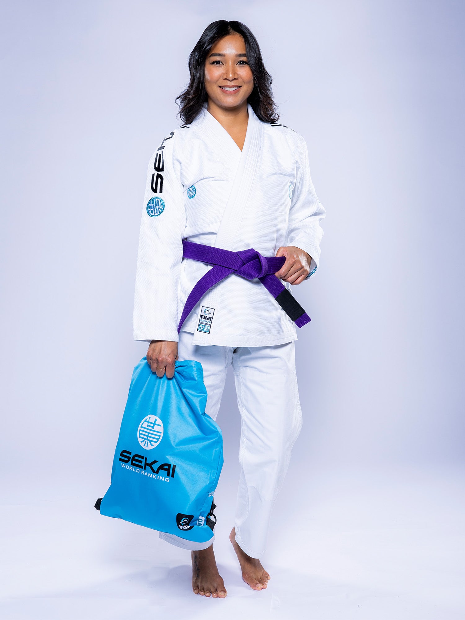 A woman in a white and teal Womens SEKAI 3.0 BJJ Gi with a purple belt stands barefoot, smiling, and holds a blue Sekai sports bag against a plain light background.