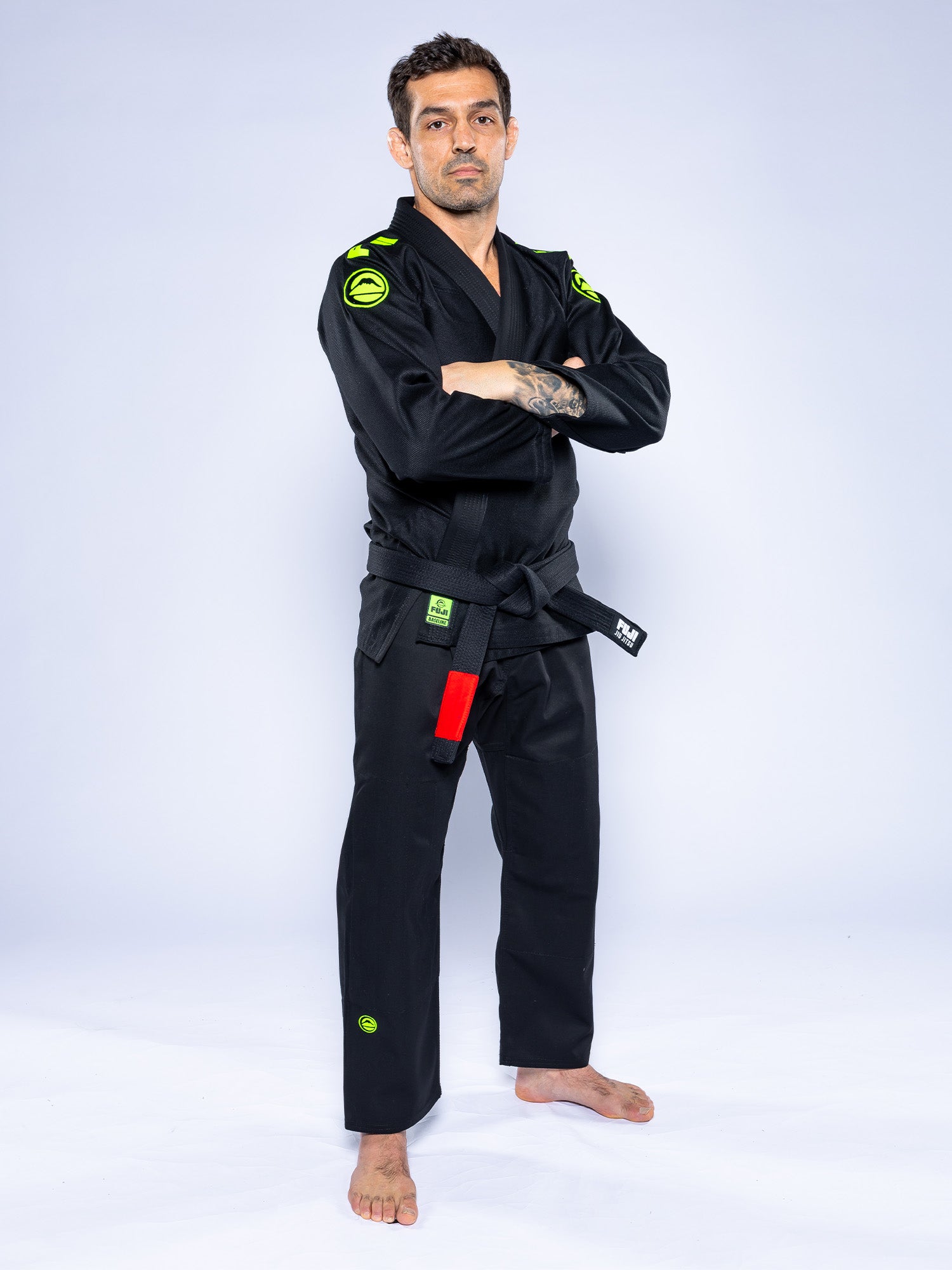 A man with short hair and left forearm tattoos stands barefoot on a light background, arms crossed confidently, wearing the Baseline 2.0 Slim Fit BJJ Gi Black Green with a black belt and green patches.