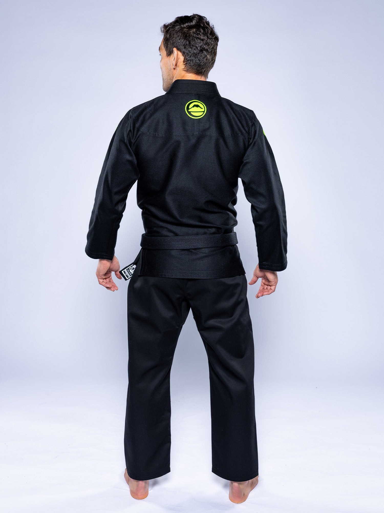 A man stands barefoot facing away, wearing the Baseline 2.0 Slim Fit BJJ Gi Black Green with a green circular logo on the upper back, against a plain light background.