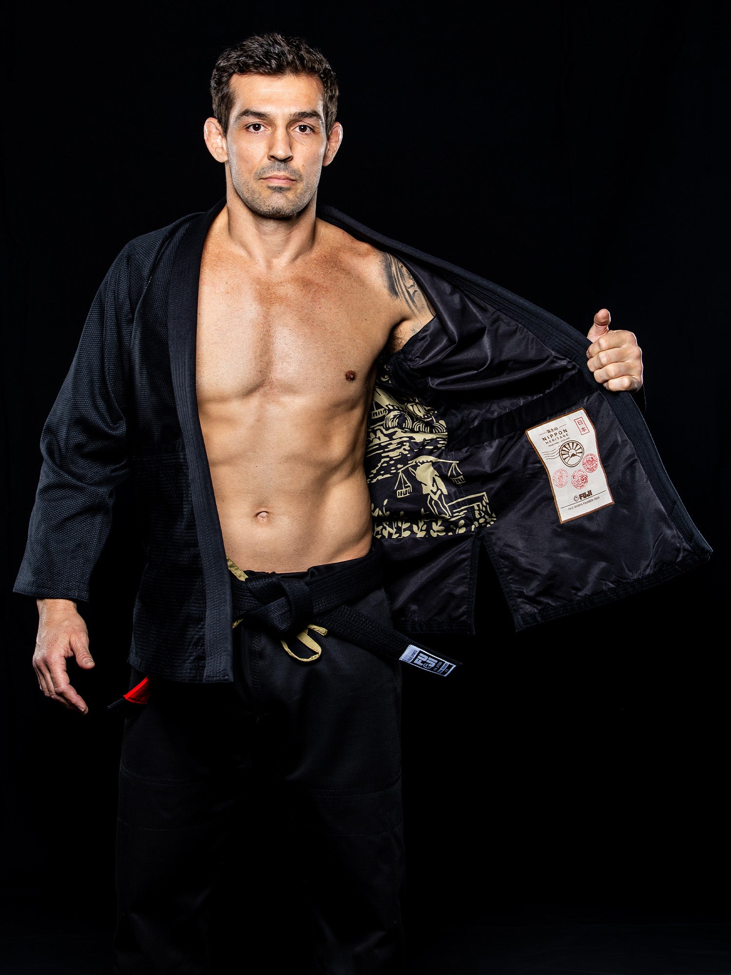 A man wearing the Heritage Keisho Lightweight BJJ Gi in black, complete with a black belt, opens his jacket to reveal its decorative inner lining while standing against a black background.