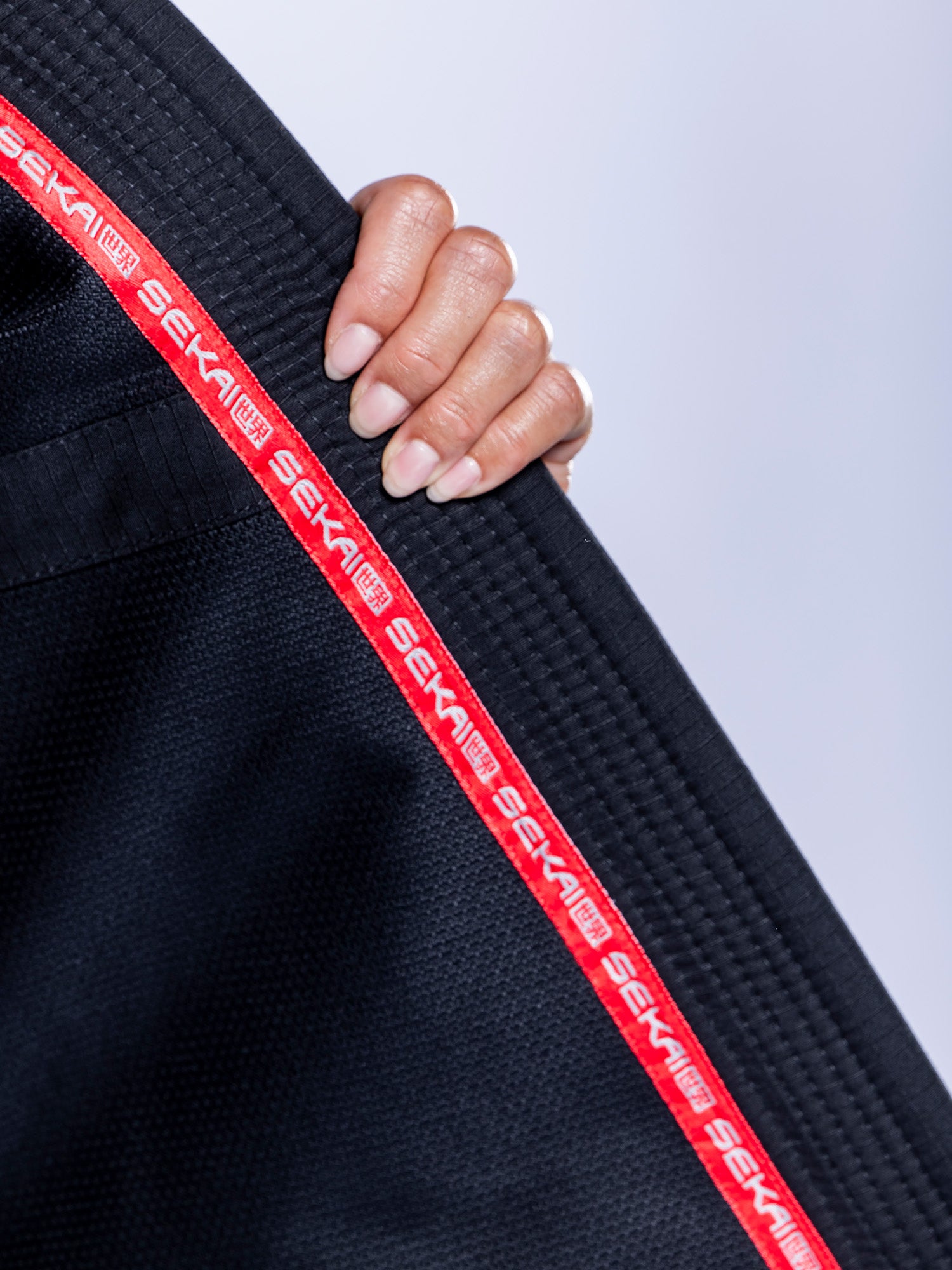 A hand holds the collar of the Womens SEKAI 3.0 BJJ Gi Black Grey, featuring black fabric with red trim and "SEKAI" repeatedly printed in white. The background is a soft, light gray.