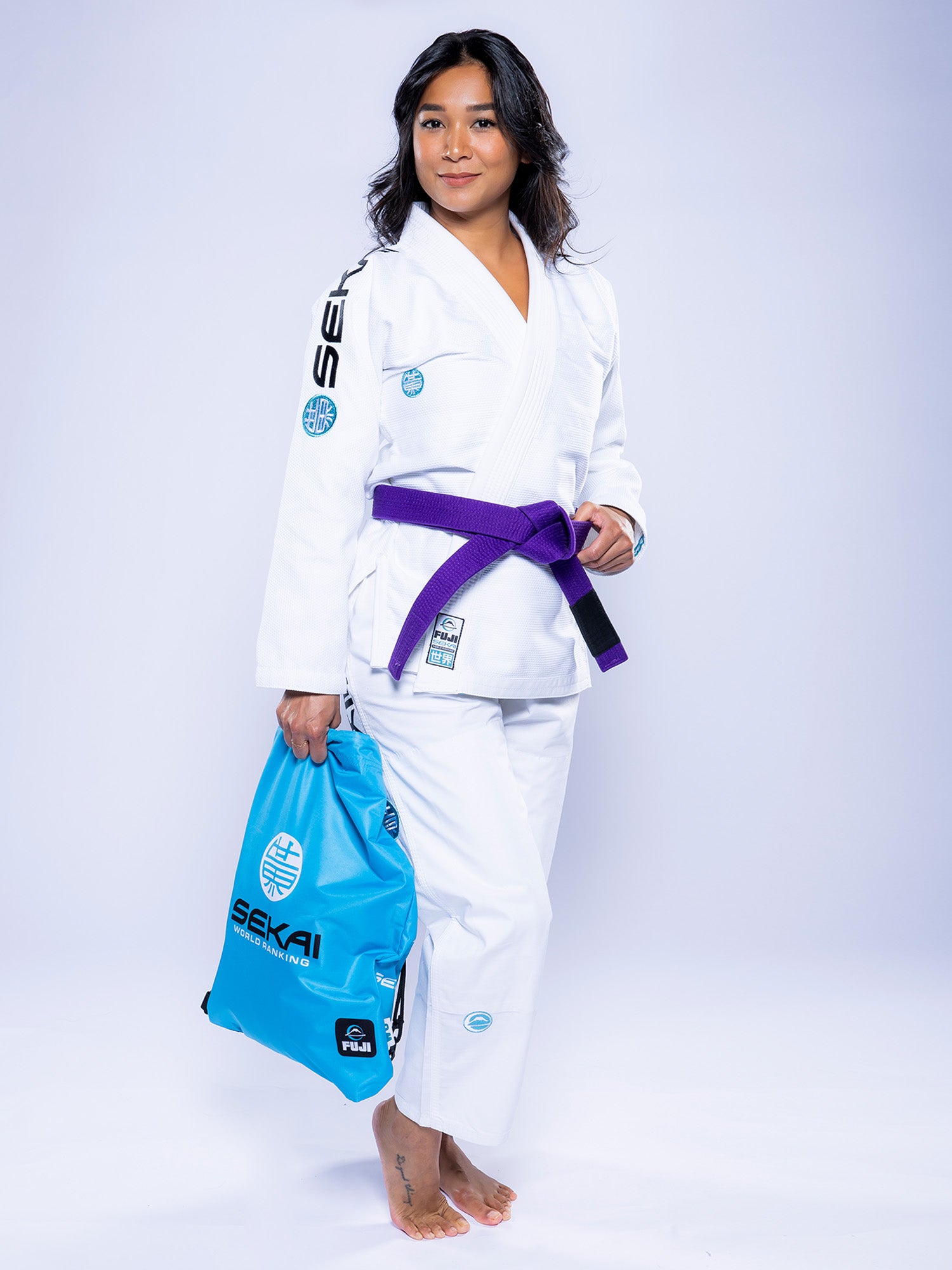 A woman wearing the Womens SEKAI 3.0 BJJ Gi White Teal with a purple belt stands barefoot on a light background, confidently smiling at the camera while holding a light blue bag.