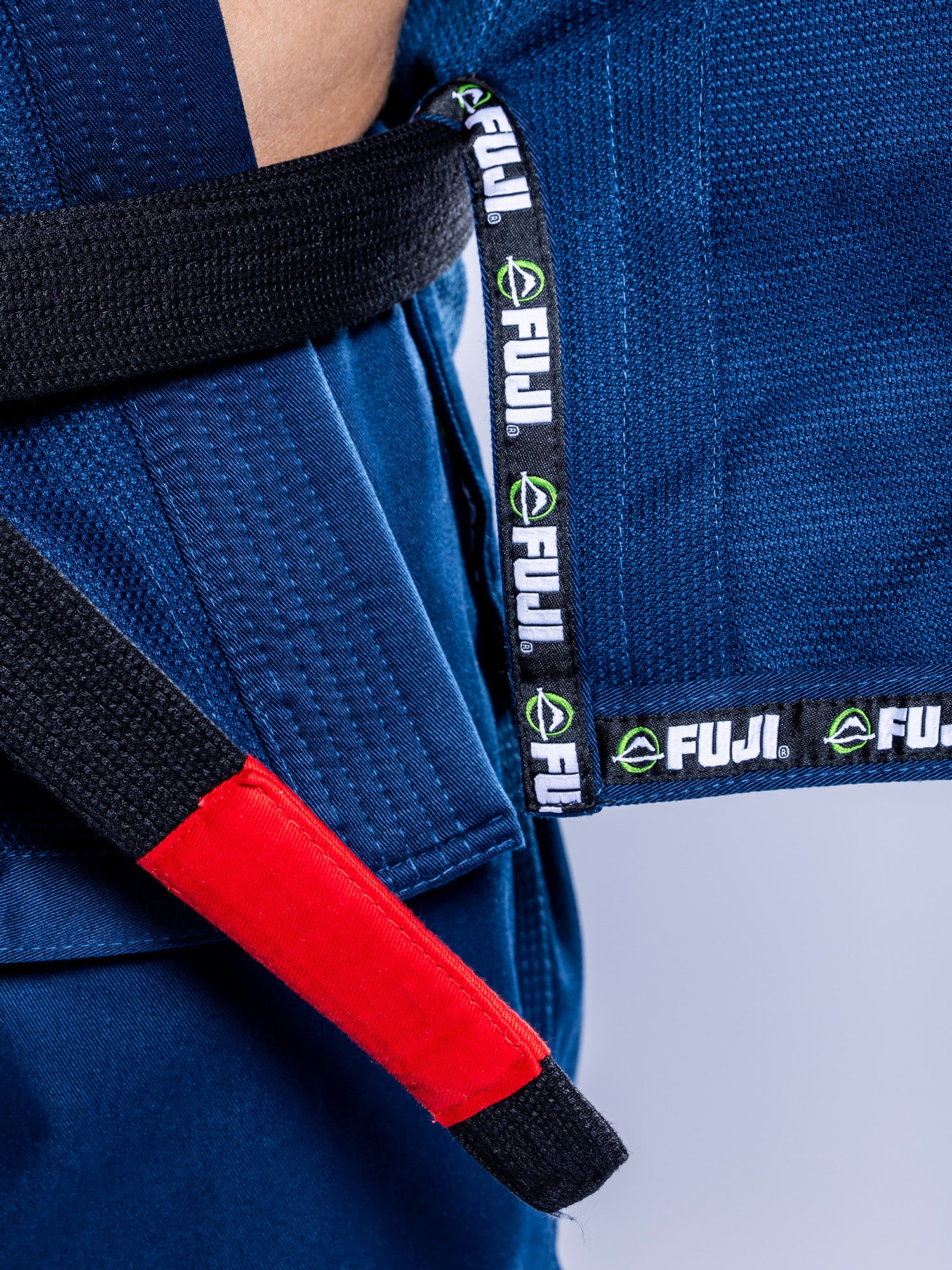 Close-up of a person wearing the Lightweight BJJ Gi Navy with a black belt featuring a red bar. The gi’s trim displays repeated FUJI brand labels.