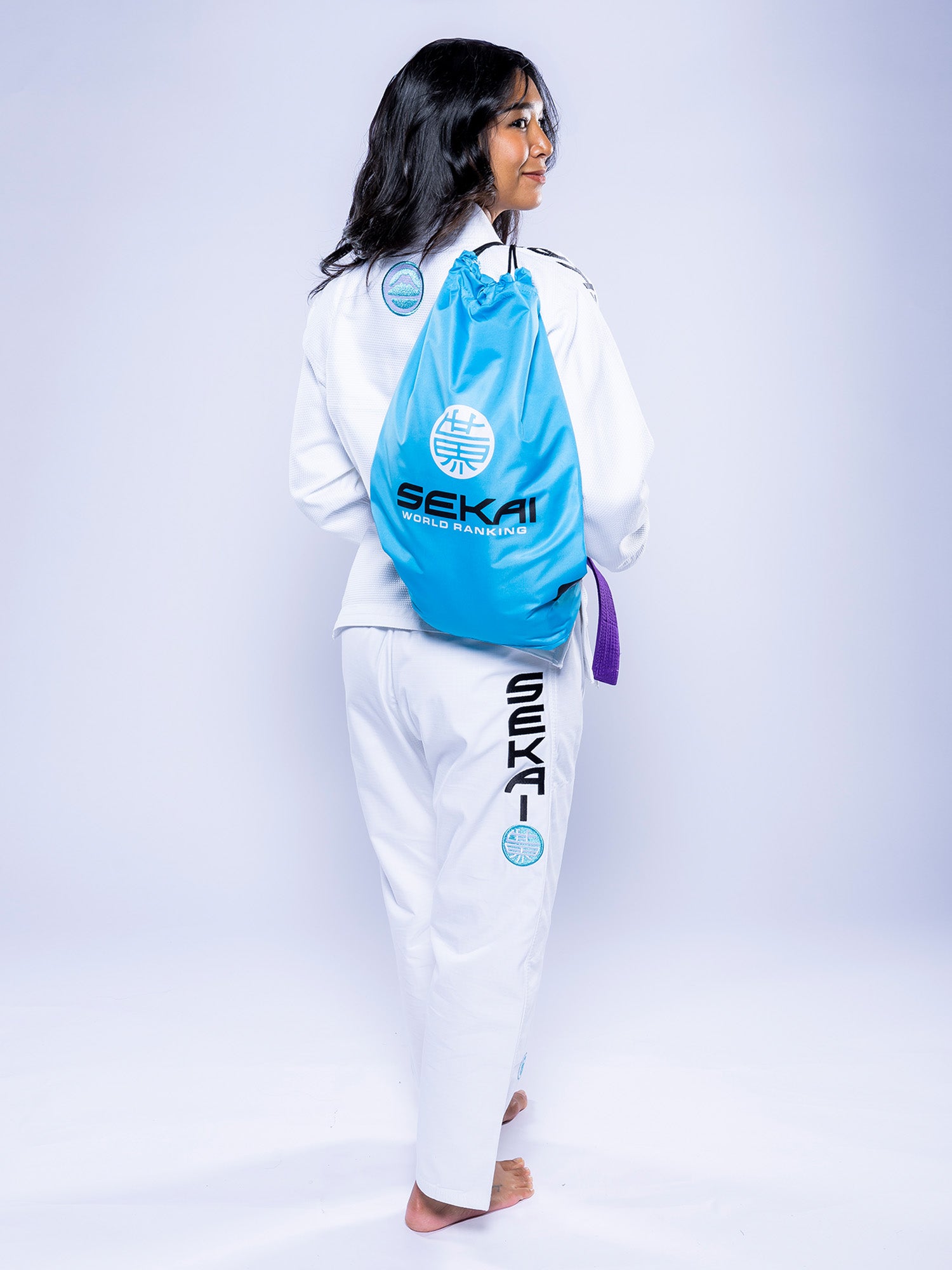 A woman in a white Womens SEKAI 3.0 BJJ Gi with teal accents and a purple belt stands barefoot, carrying a bright blue “SEKAI” logo drawstring bag. The background is plain and light.