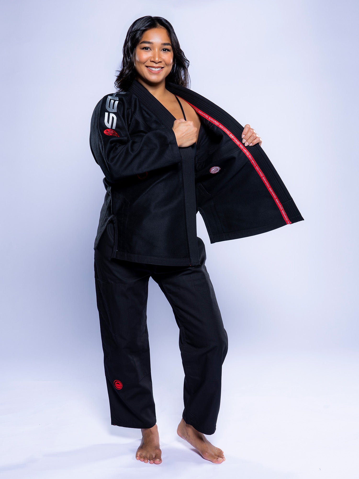 A woman smiles at the camera, barefoot in the Womens SEKAI 3.0 BJJ Gi Black Grey, holding open her jacket to display the inner lining against a plain light gray background.