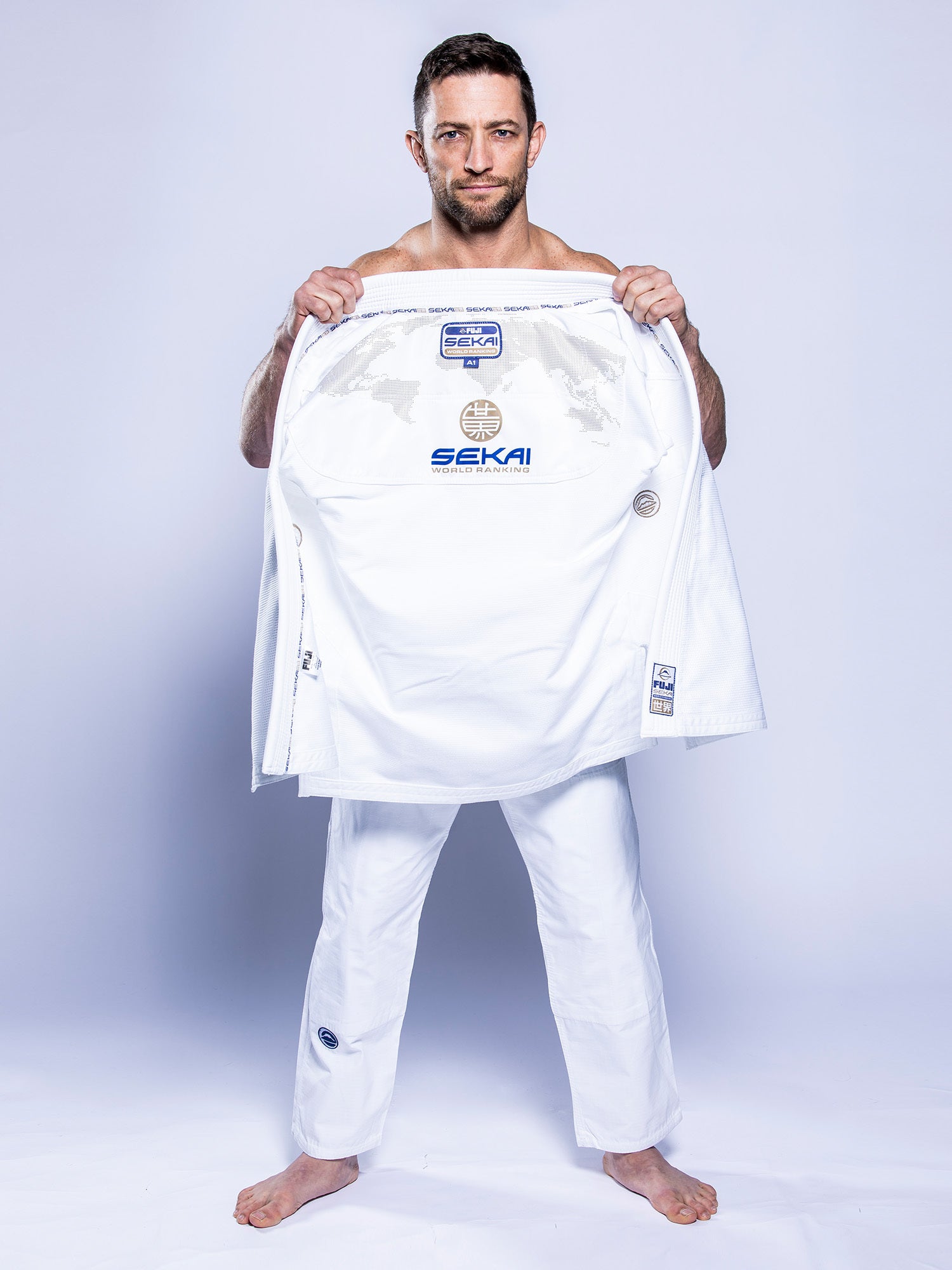 A man stands barefoot on a white floor, holding open his white SEKAI 3.0 BJJ Gi (White Navy) jacket to reveal the inside with visible “Sekai” branding. He looks at the camera against a plain light gray background.