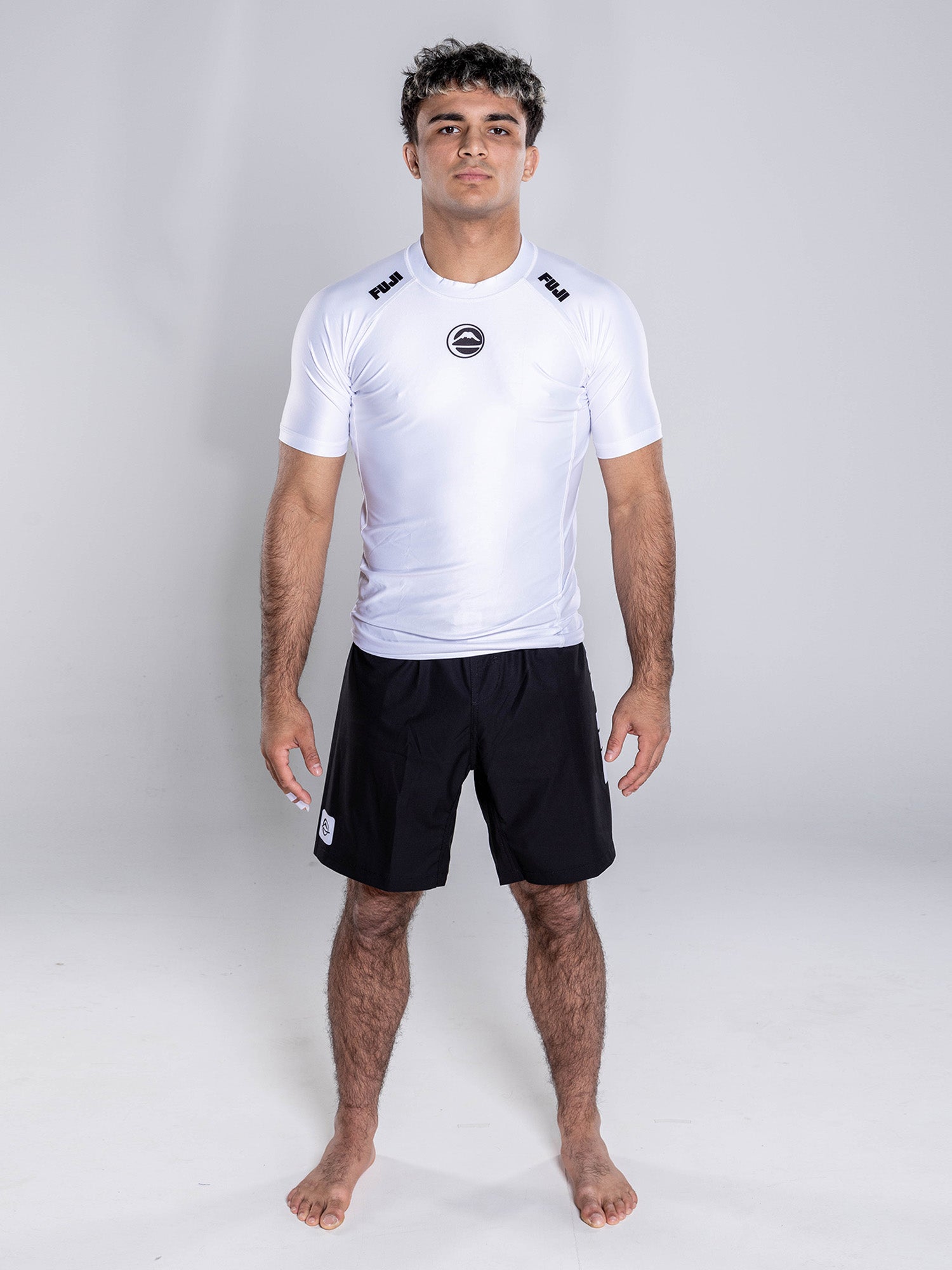 A barefoot person stands facing forward against a plain background, wearing the Baseline 2.0 Short Sleeve Rashguard White and black shorts.