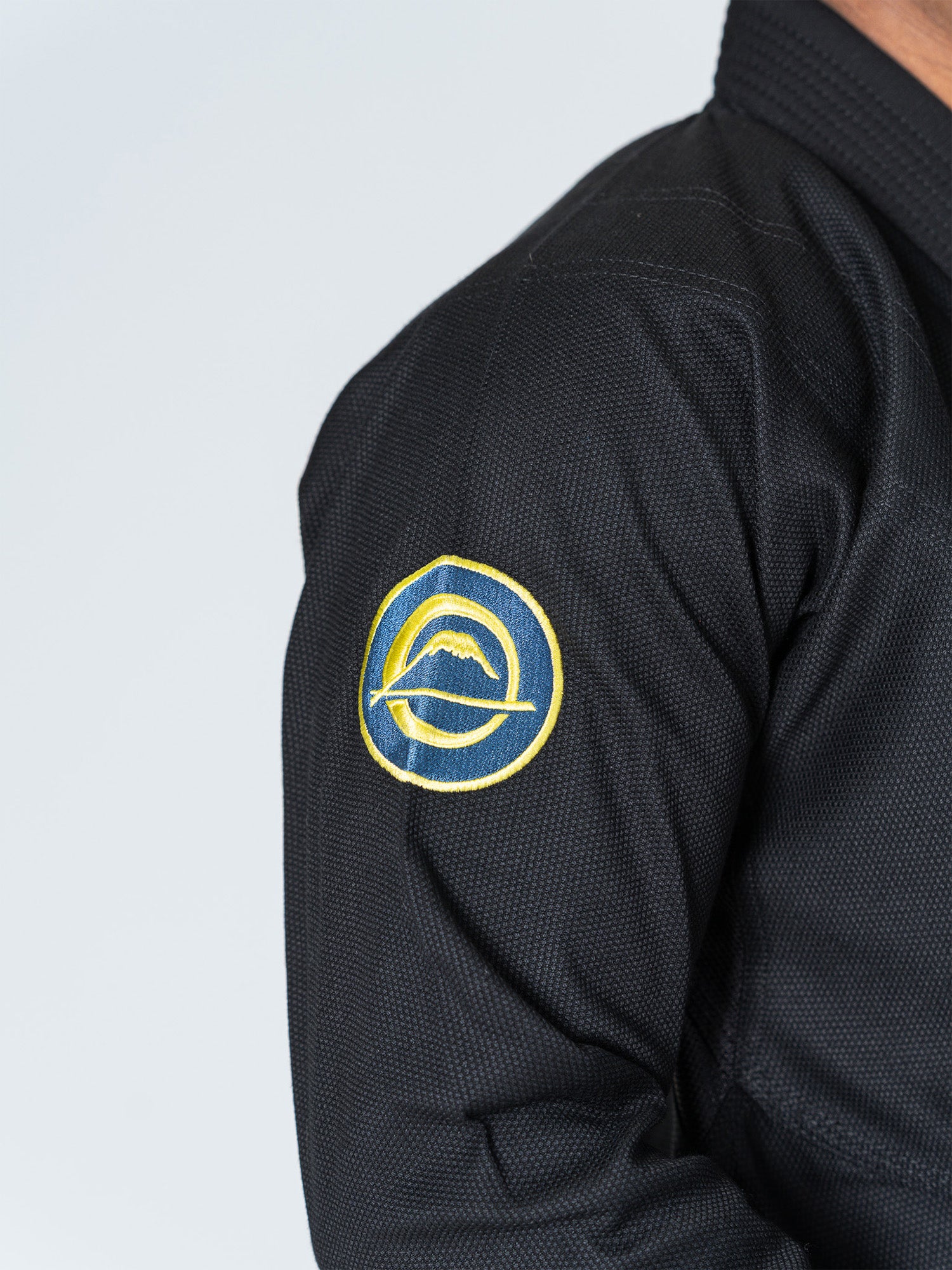 Close-up of a person wearing the Special Edition Holiday Flow Tech Jiu Jitsu Gi Black, featuring a circular yellow and blue patch with a wave design on the upper sleeve.