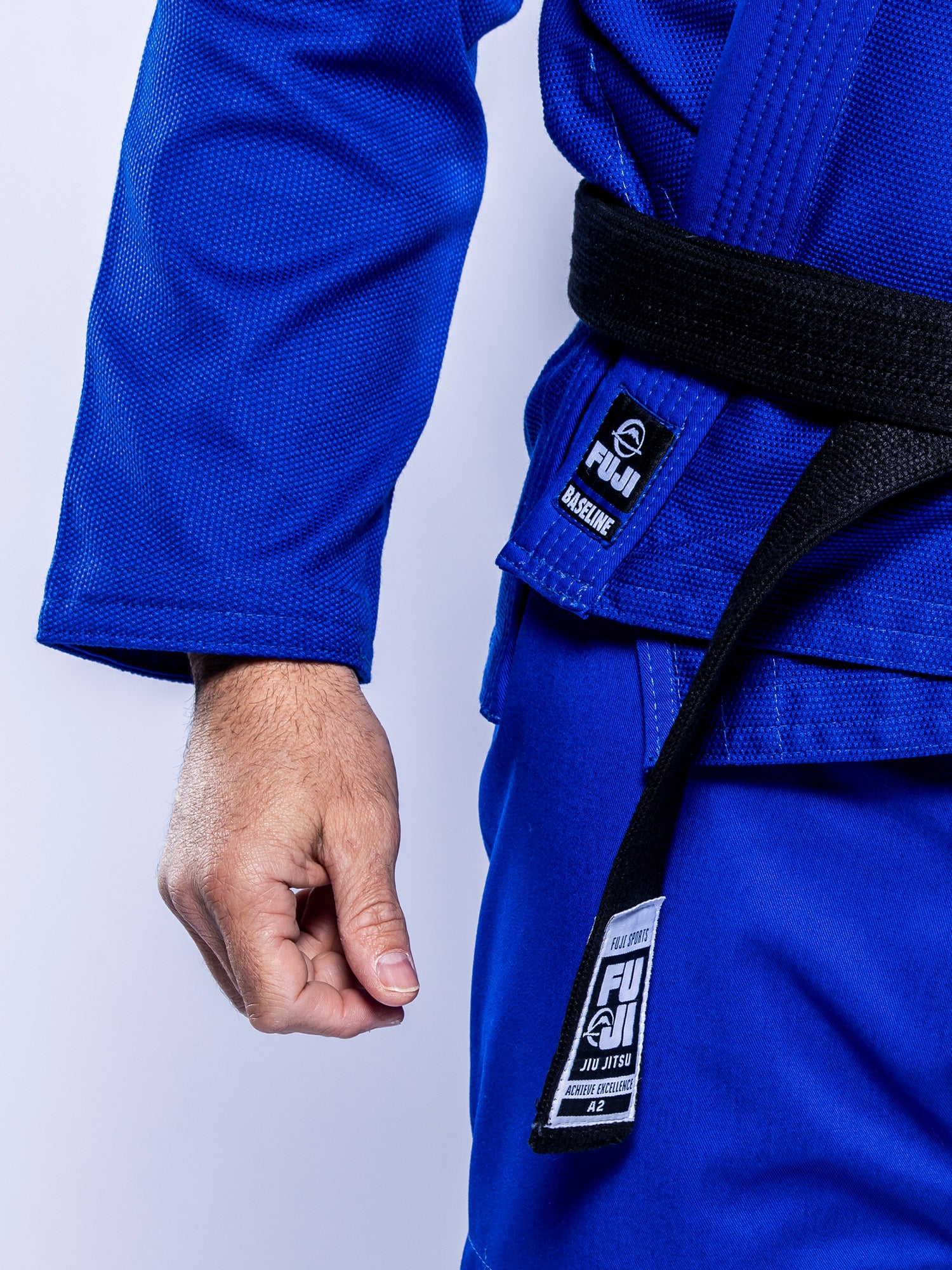 A person in the Baseline 2.0 BJJ Gi Blue White with a black belt stands with a clenched fist, showing visible Fuji branding tags.